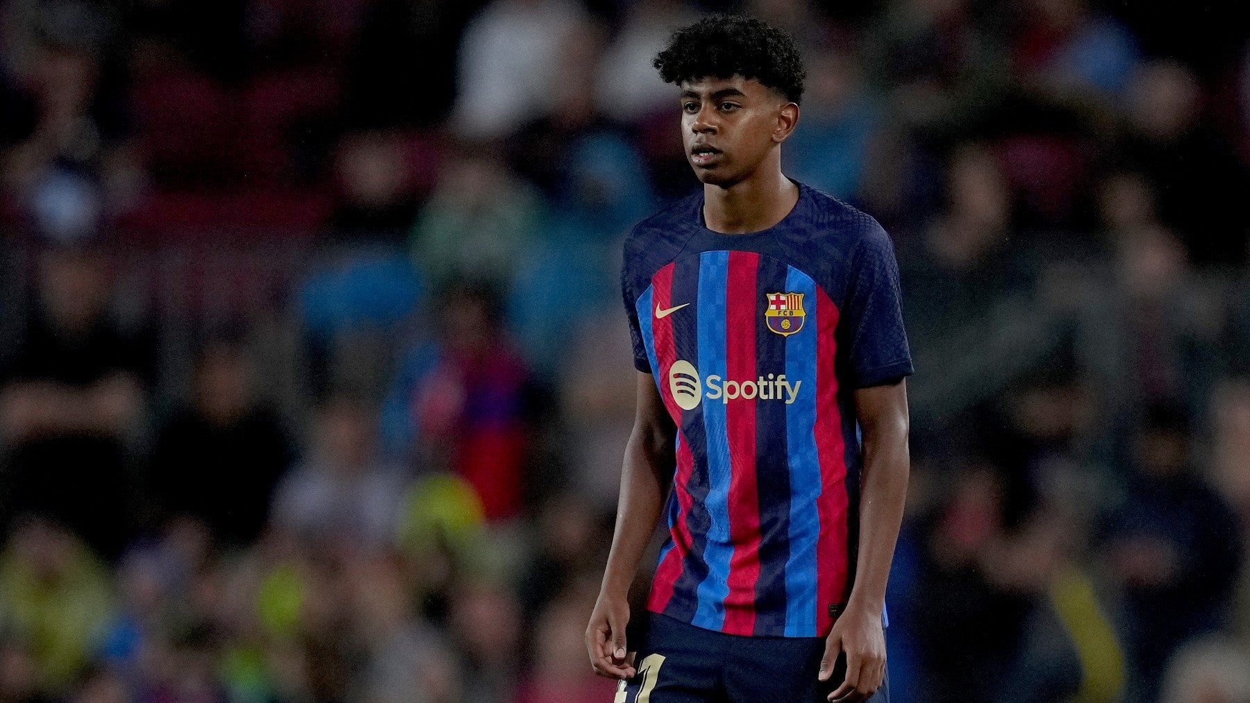 Lamine Yamal of FC Barcelona in action during a match. He wears the team's signature blue and red jersey with the Spotify logo on the front. The background shows a blurred stadium crowd
