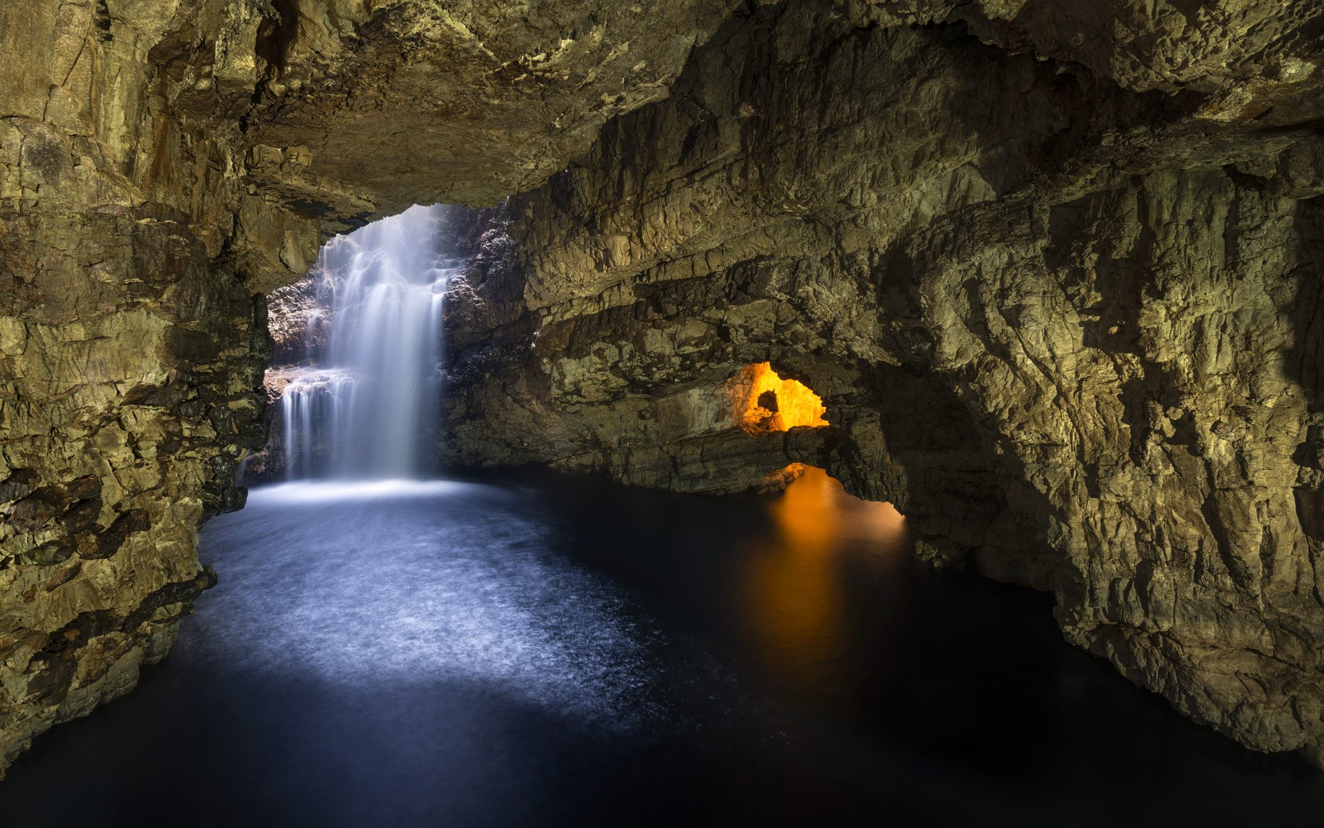 A mesmerizing long exposure photograph of a waterfall cascading into a subterranean pool within a cave. The cave walls are illuminated with warm, inviting light, and the water appears smooth and ethereal