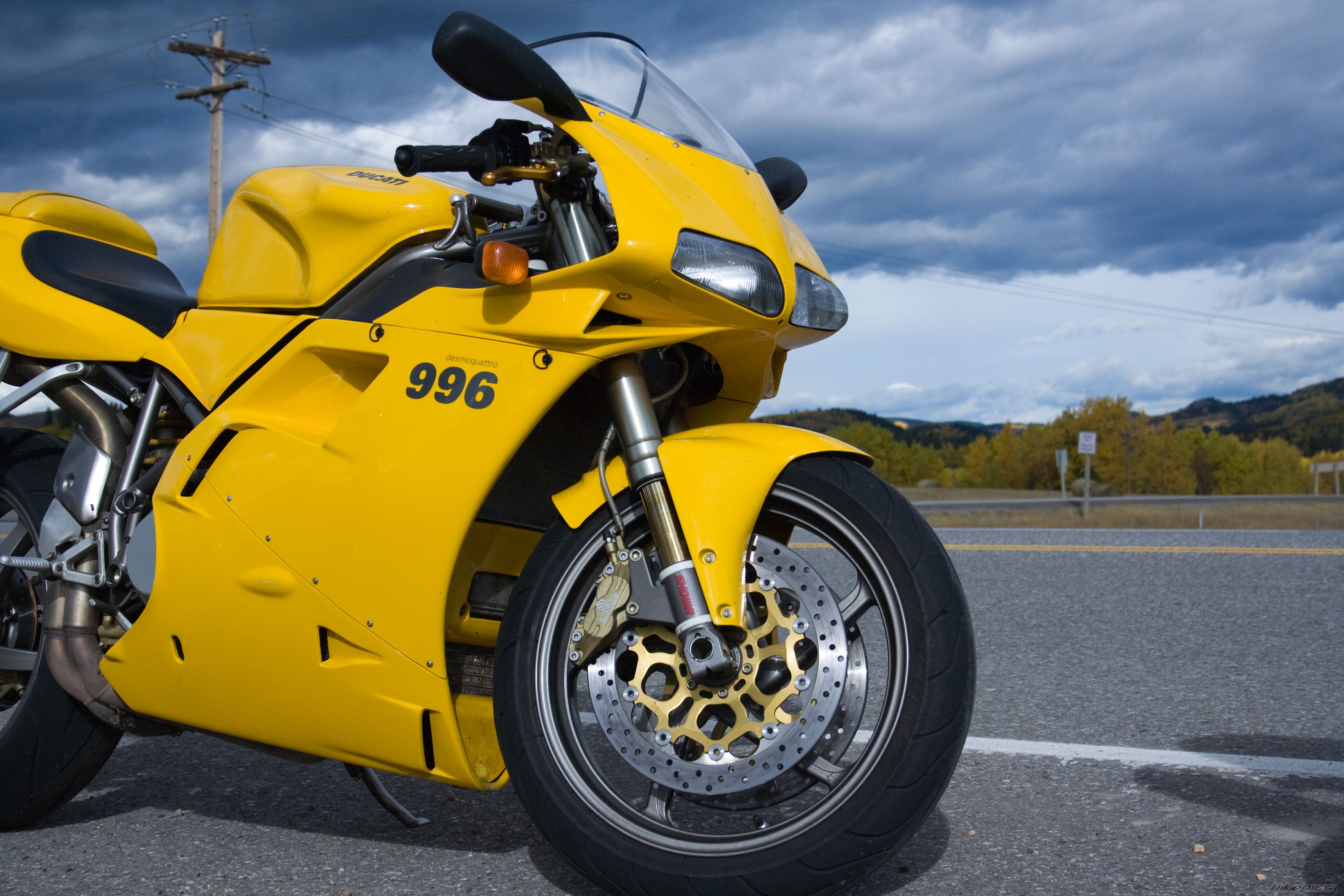 A vibrant yellow Ducati 996 motorcycle parked on the side of a road, set against a backdrop of a dramatic cloudy sky and autumnal trees