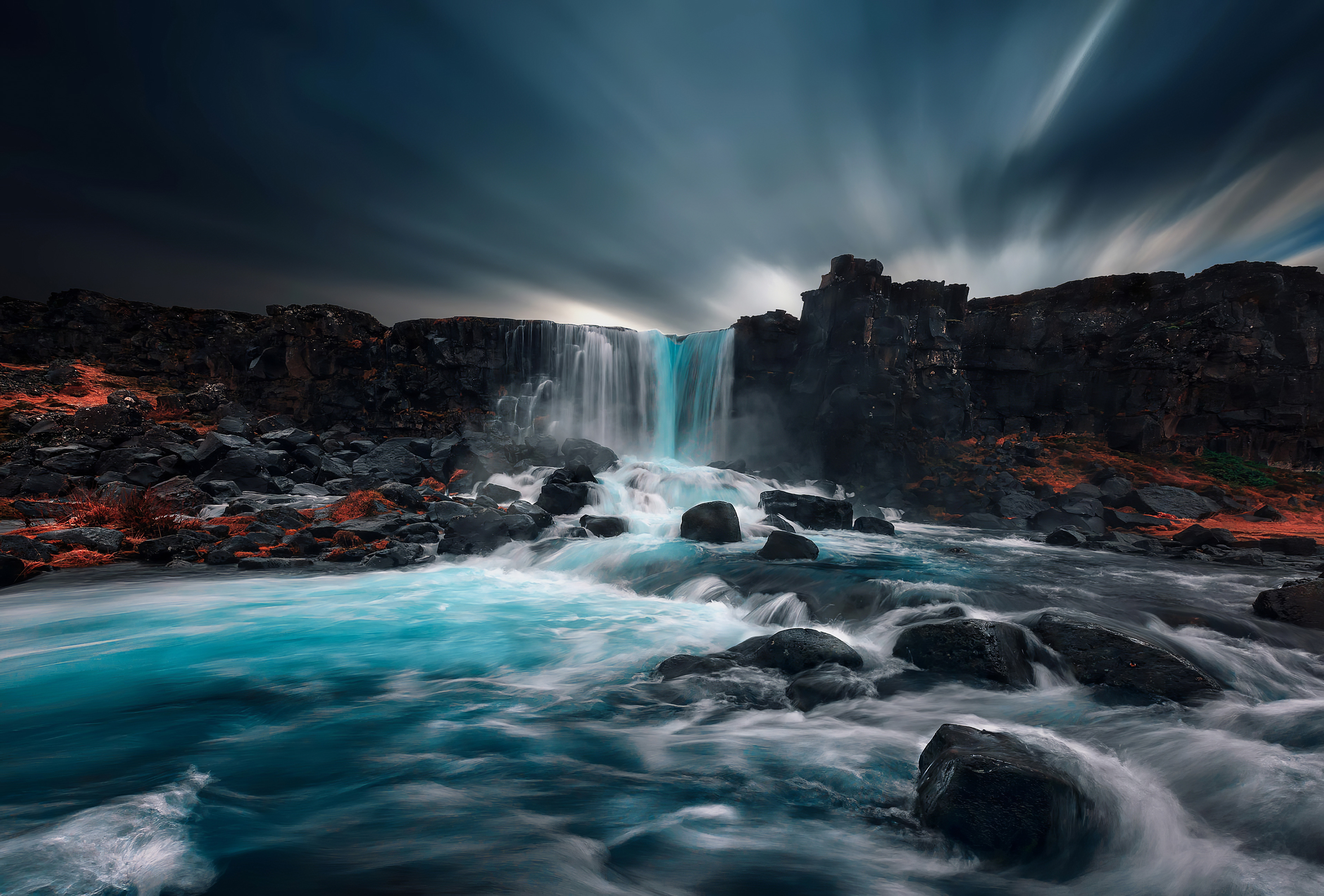 A stunning 4k wallpaper of a waterfall cascading over dark rocks into a vibrant turquoise river. The long exposure photography captures the smooth flow of the water and dramatic movement of the clouds