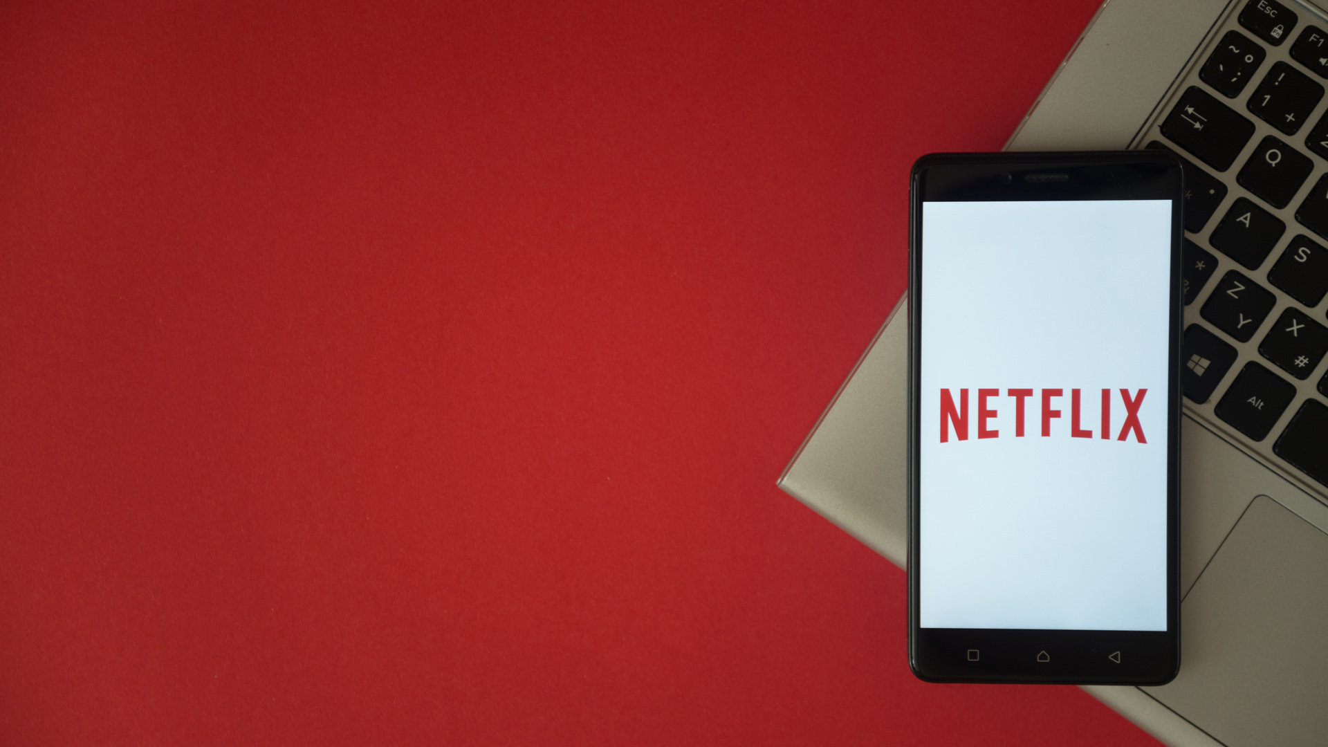 Netflix logo displayed on a smartphone screen, placed on a laptop keyboard, with a red background