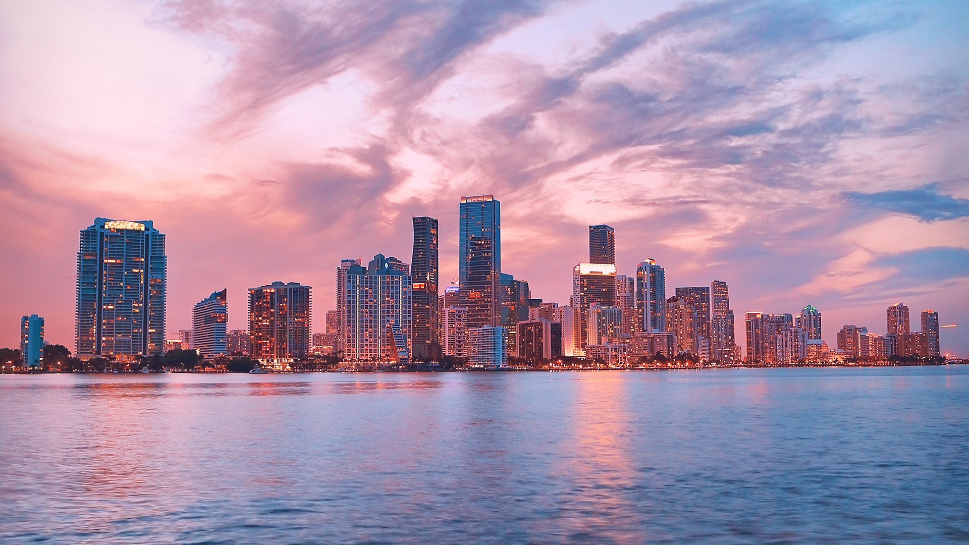 A stunning sunset cityscape of Miami, Florida, with vibrant pink and purple hues reflecting on the calm waters. The Miami skyline is prominently featured, showcasing the city's modern architecture and vibrant urban landscape