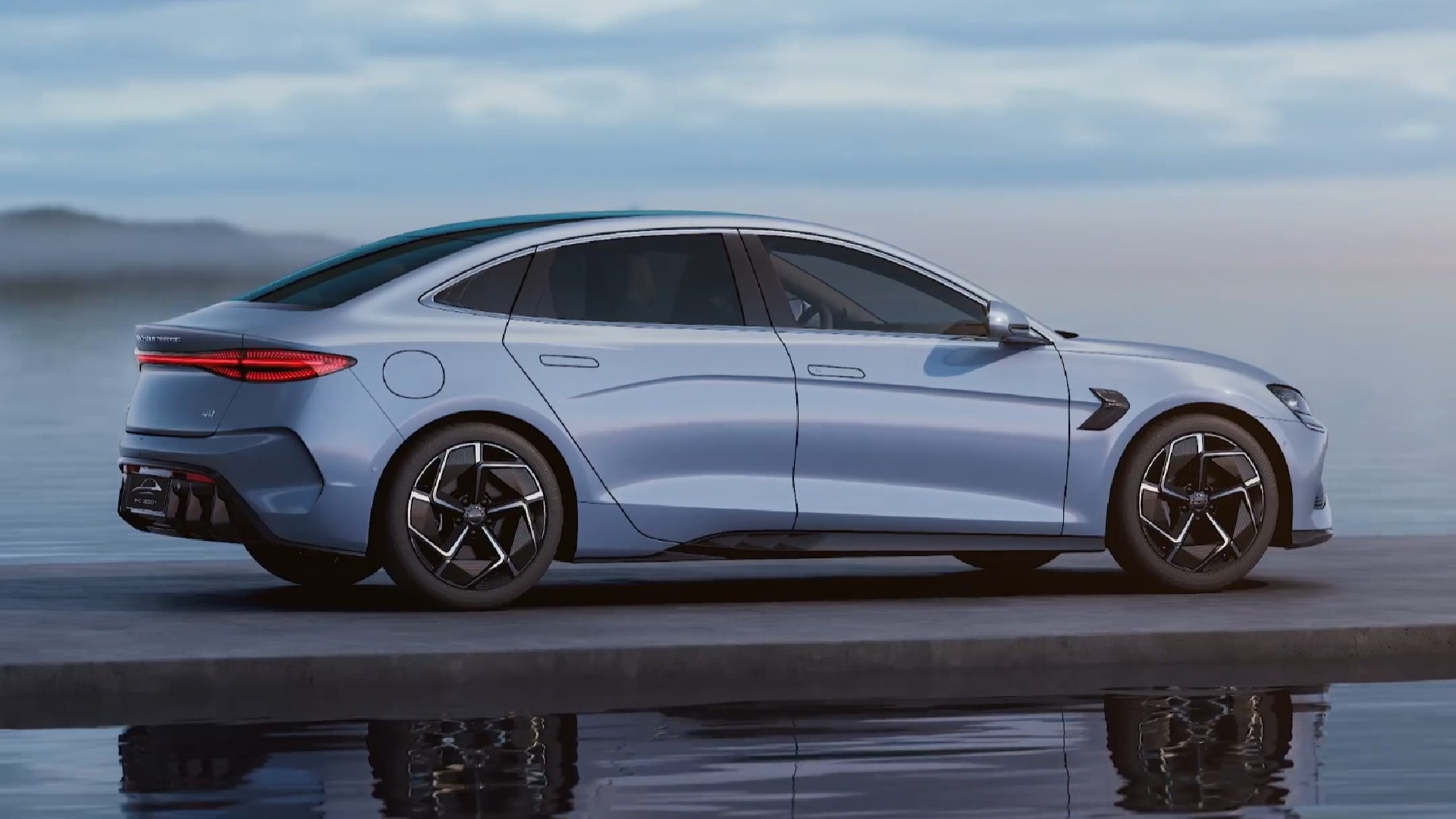A sleek, silver BYD electric sedan parked by the water, reflecting the serene sky and surrounding landscape