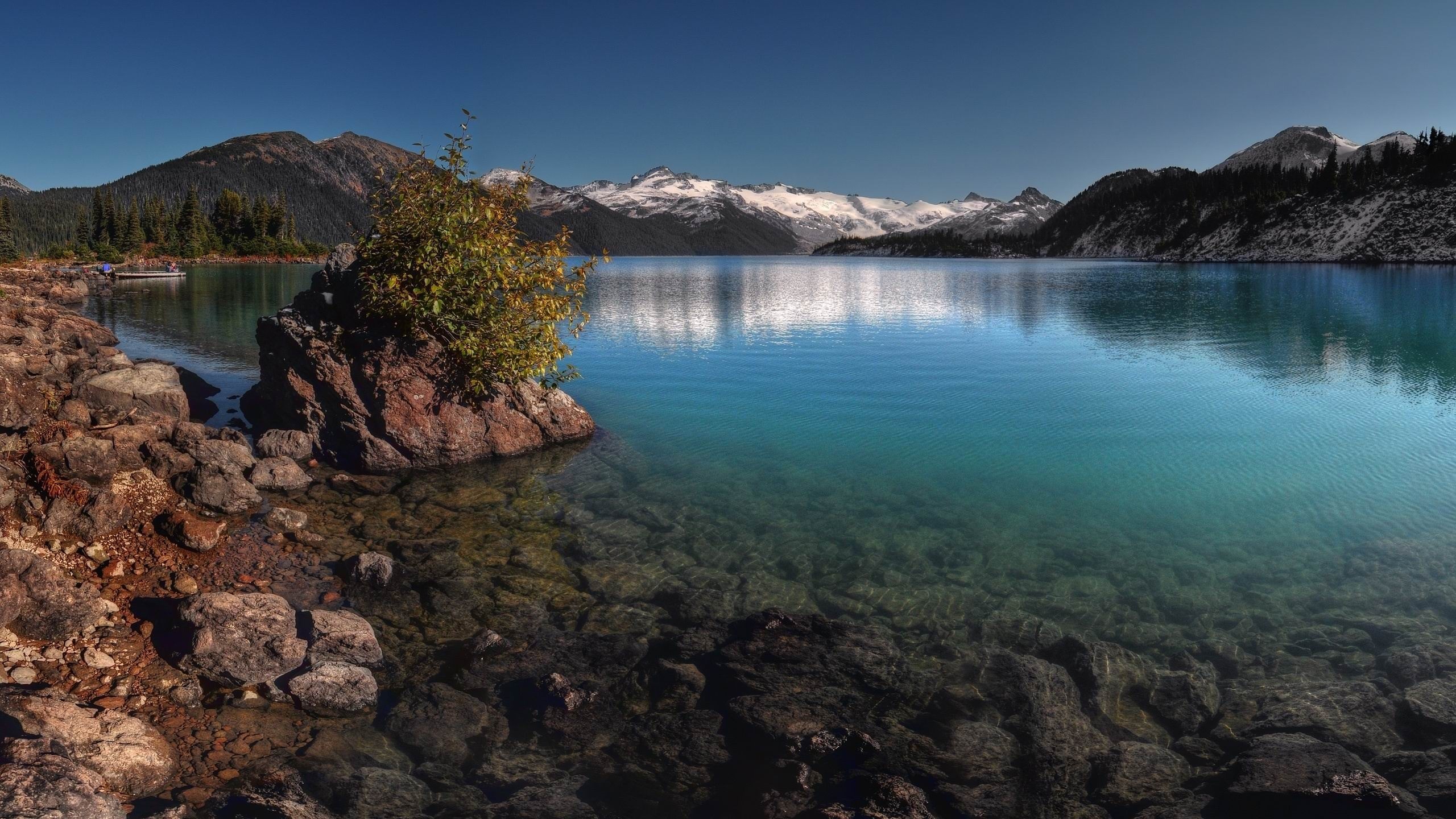 A breathtaking landscape photograph showcasing a pristine alpine lake. The crystal-clear turquoise water reveals the rocky bottom. A small, rocky island dotted with shrubs anchors the foreground, while snow-capped mountains rise majestically in the background