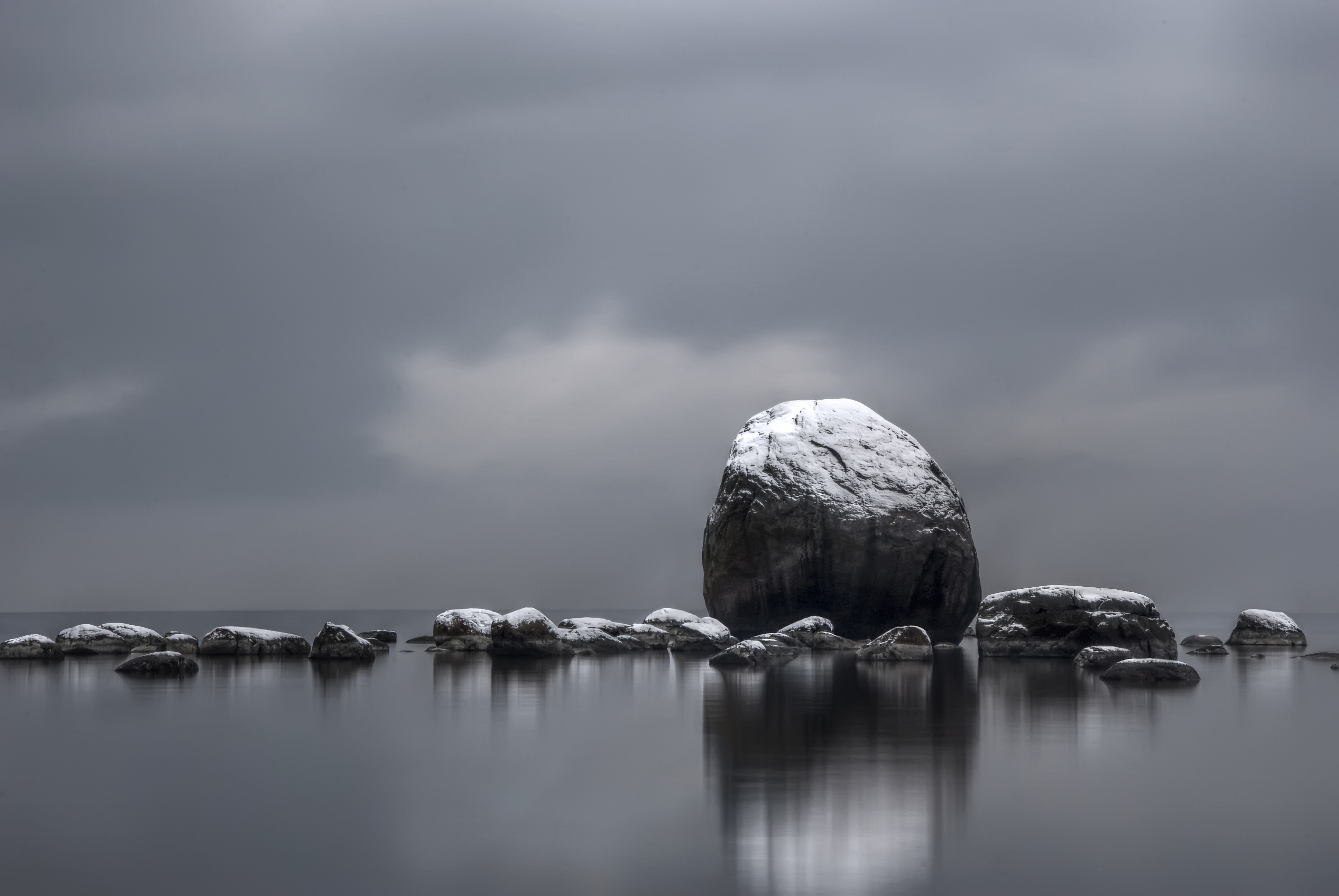 A serene 4K wallpaper featuring a snow-dusted boulder rising from the calm waters of a lake under a moody, cloud-filled sky