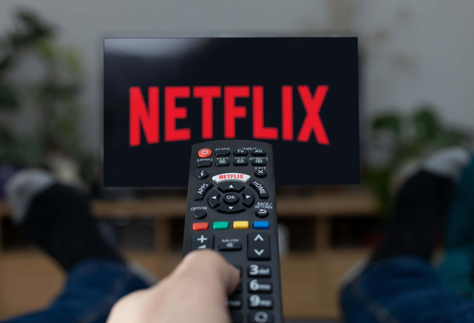 A person relaxes on a couch, feet up, using a remote to navigate the Netflix app on their TV. The iconic Netflix logo is prominently displayed on the screen