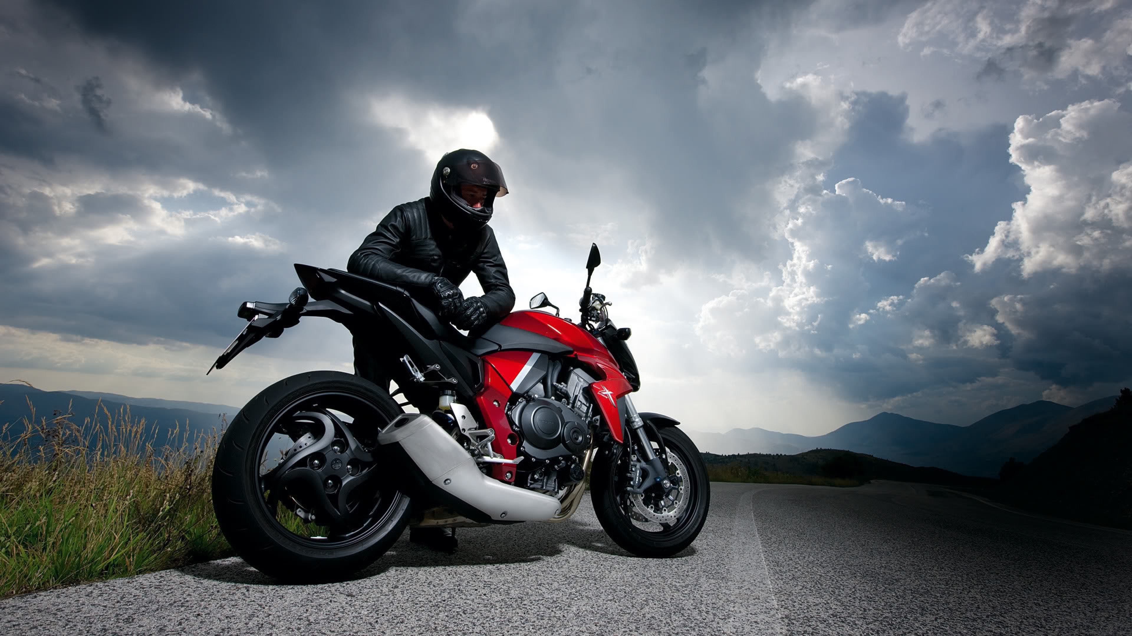 A motorcyclist pauses on a mountain road, taking in the dramatic view under a cloudy sky