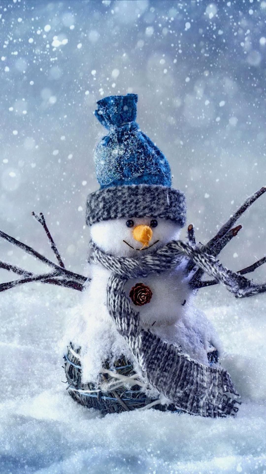 A charming snowman figurine sits amidst a snowy scene, adorned with a blue hat, a gray scarf, and twig arms.  The backdrop features gently falling snow, adding to the winter wonderland ambiance