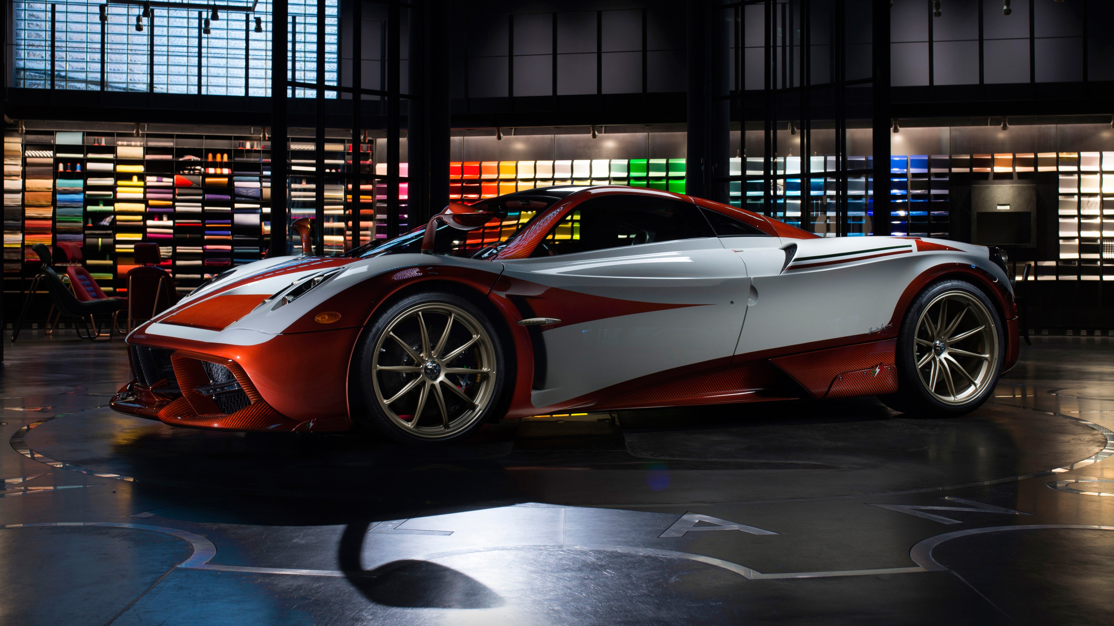 A stunning Pagani Huayra sports car, showcasing its unique two-tone paint job and sleek design, parked in a luxurious showroom with a colorful wall of leather samples in the background
