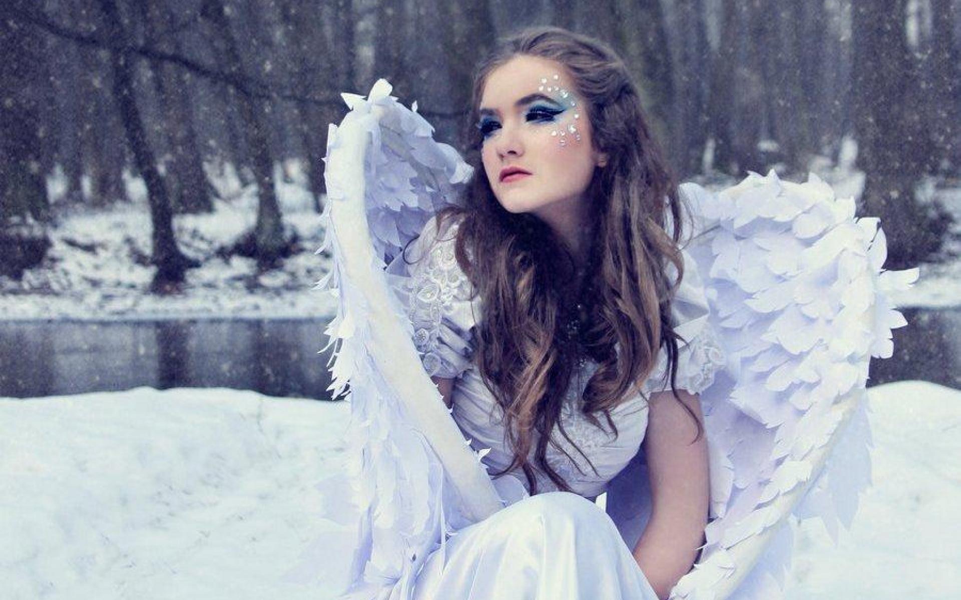 A woman dressed as a snow angel sits in a snowy forest, her white wings spread out behind her.  She wears a white dress and has a serene expression on her face