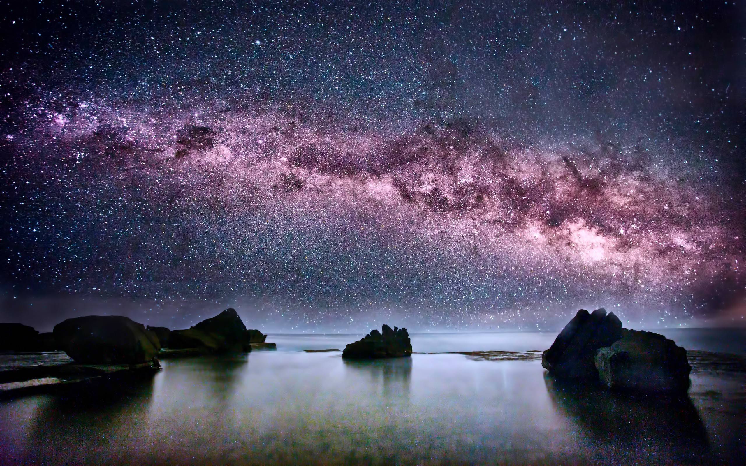 A breathtaking nightscape with the Milky Way galaxy stretching across the sky above a tranquil body of water with dark rocks emerging from the surface