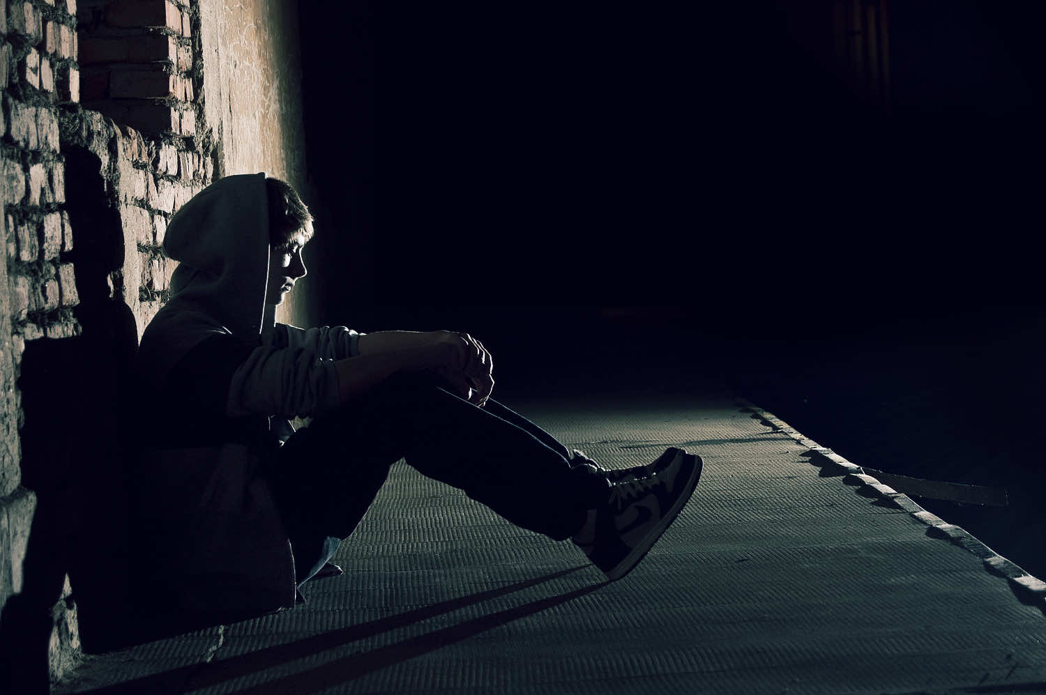 A solitary figure hunched over in the shadows against a brick wall, conveying a sense of sadness and loneliness