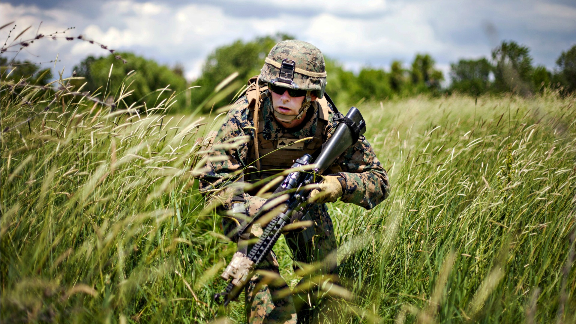 A soldier in camouflage uniform, equipped with a rifle, stealthily advances through tall grass in a military training exercise