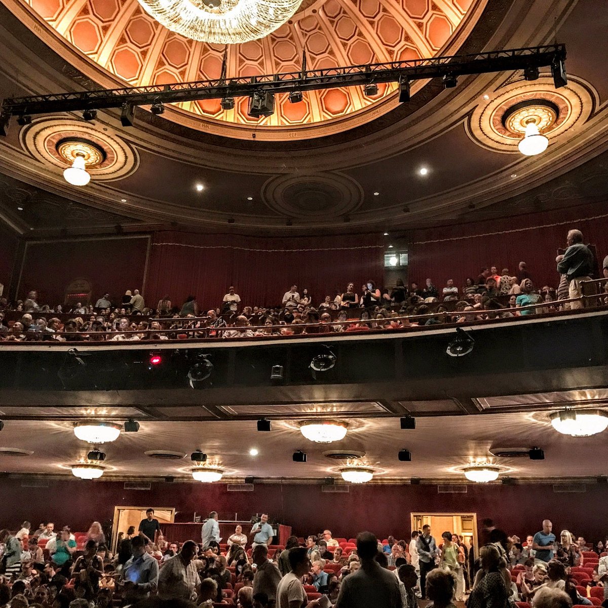 A view from the orchestra section of a Broadway theater, showing the audience in the mezzanine and the ornate ceiling