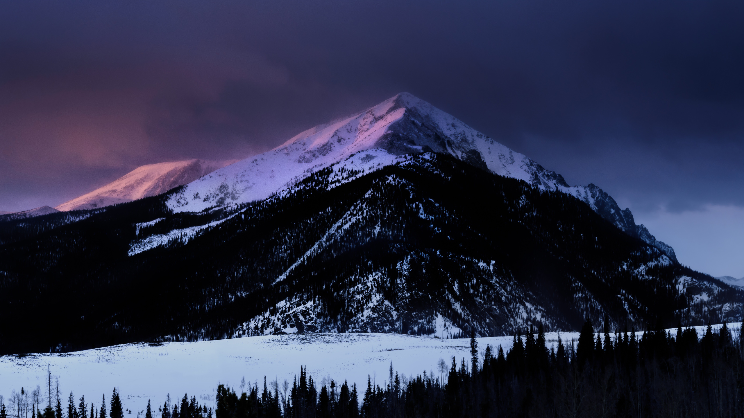 A breathtaking 4K wallpaper showcasing a snow-capped mountain peak at sunset. The mountain's slopes are covered in a blanket of fresh snow, and the sky is painted with vibrant hues of pink and purple