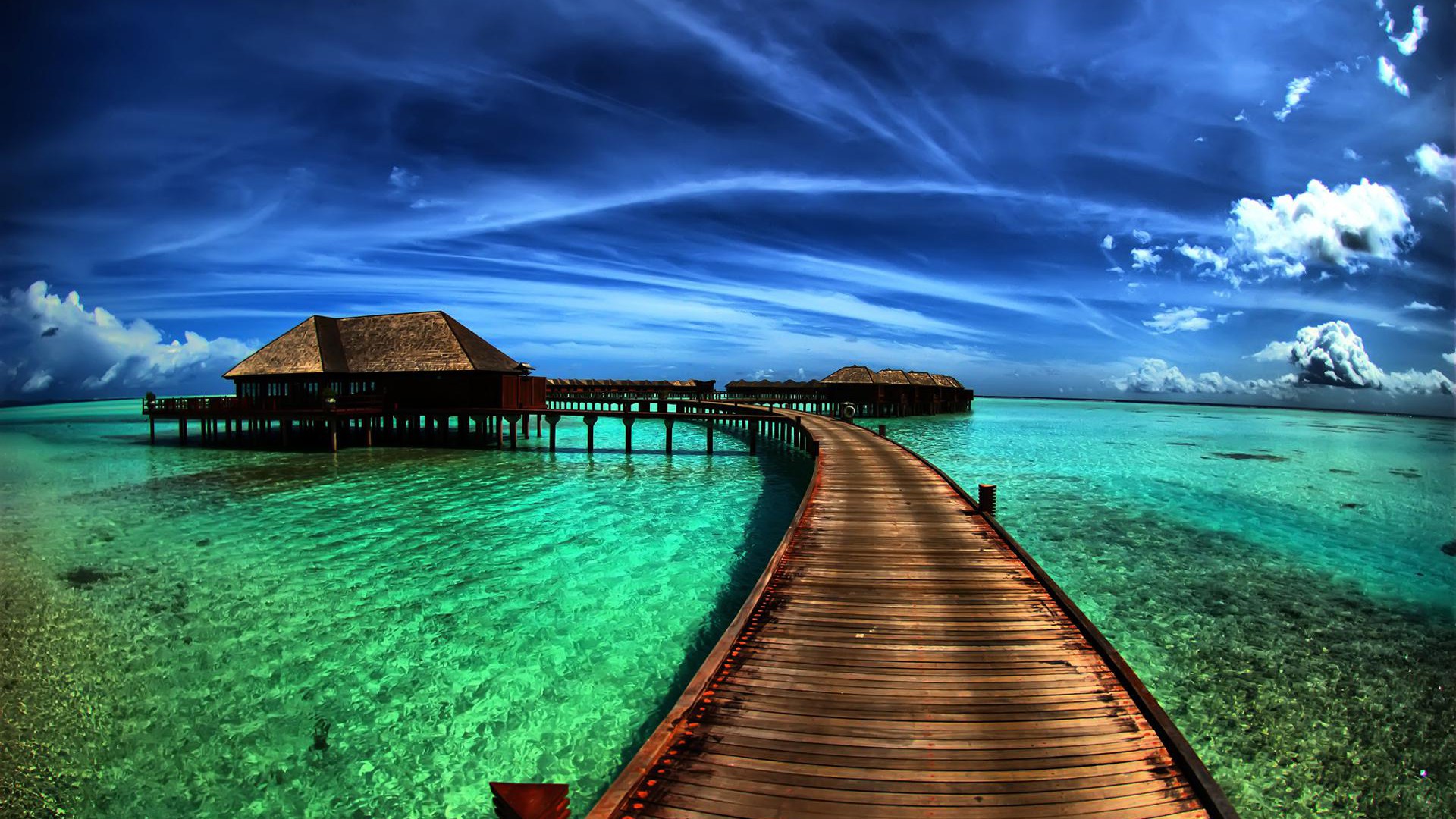 A wooden walkway leading to overwater bungalows in crystal-clear turquoise water, under a vibrant blue sky with wispy clouds
