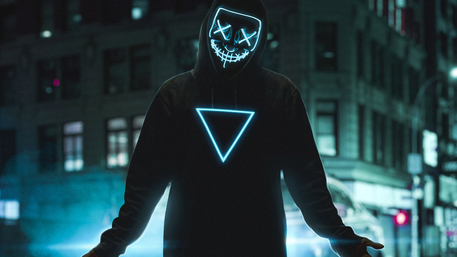 A person wearing a black hoodie and an LED mask with a glowing triangle design stands in a dark city street at night