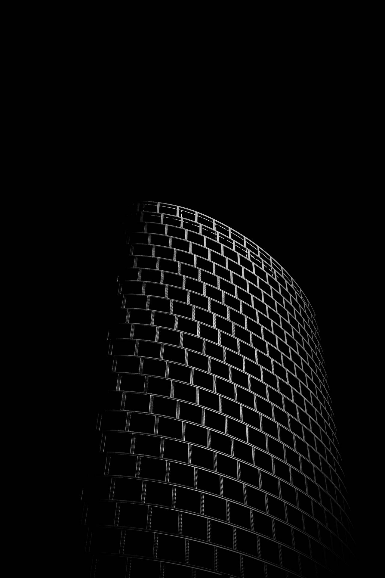 A dramatic black and white photograph of a cylindrical building's facade at night, highlighted against a dark background, showcasing its grid-like pattern of windows