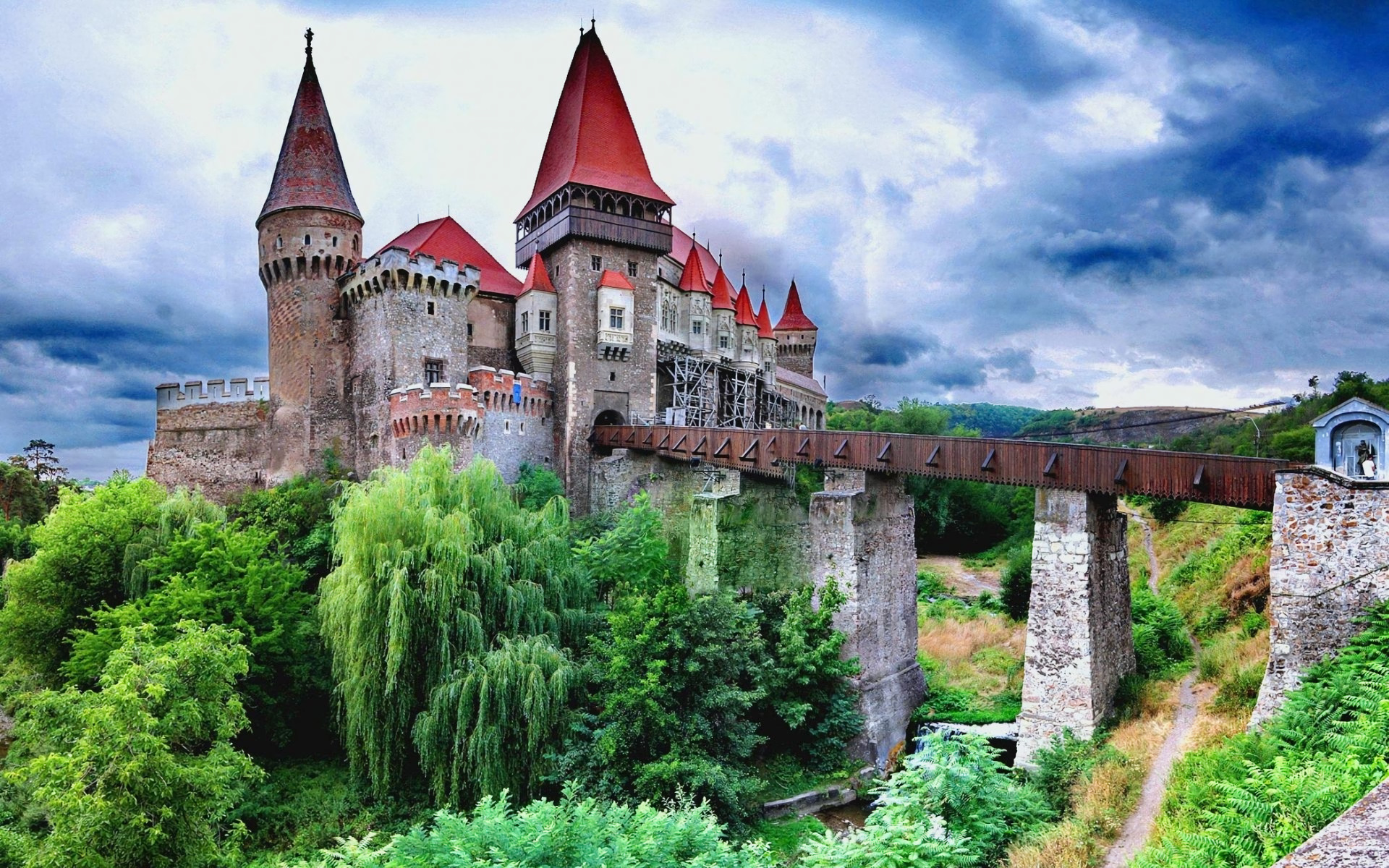 A majestic castle with red roofs and towering spires stands on a hill, connected to the foreground by a long wooden bridge. Lush green foliage surrounds the castle and the river below, creating a picturesque scene