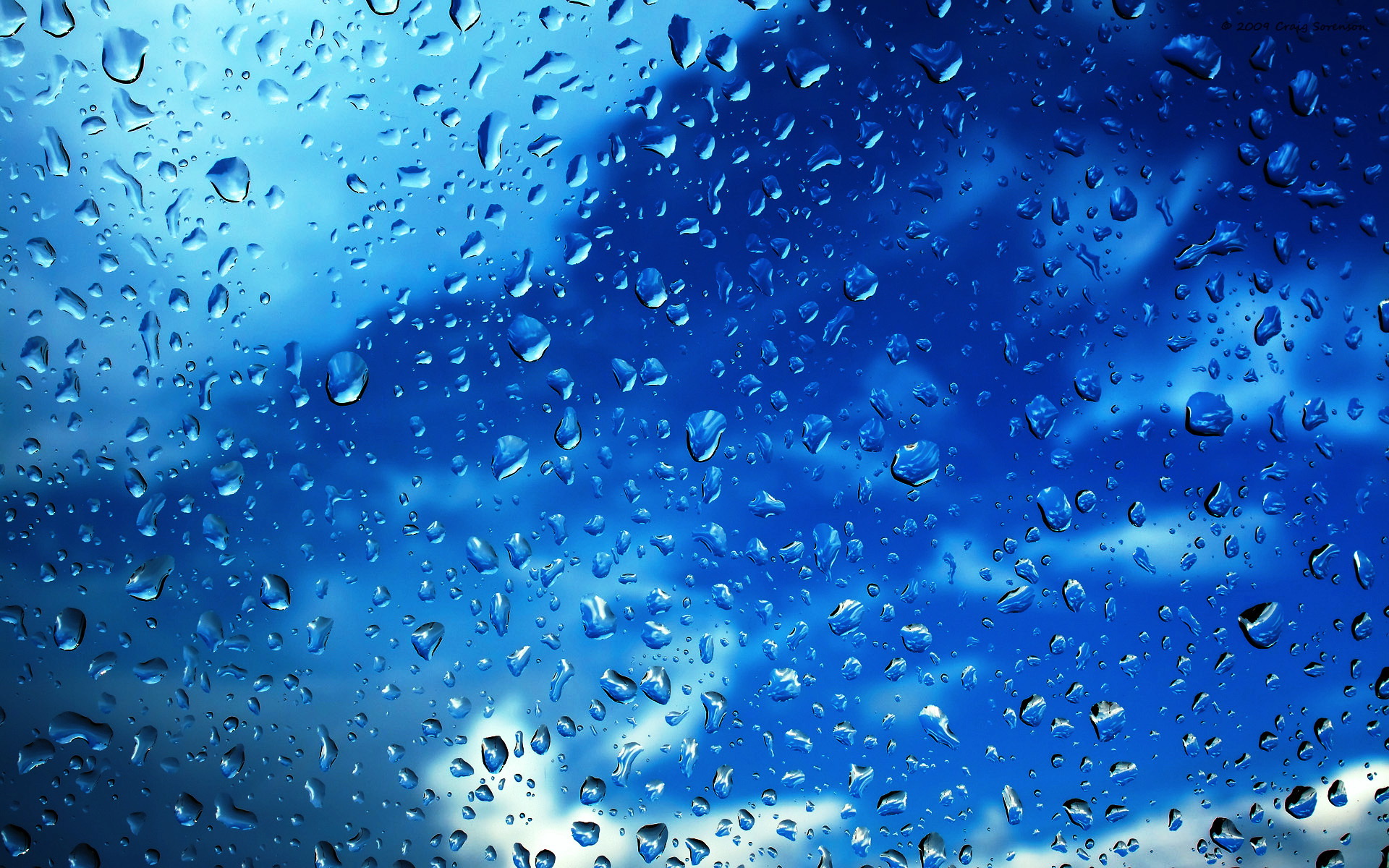 Close-up of raindrops on a window against a vibrant blue sky with soft white clouds, creating a tranquil and refreshing scene