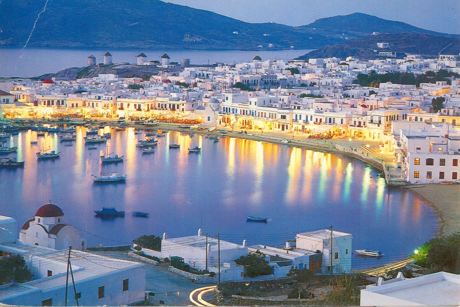 Mykonos Town, Greece, at dusk. Whitewashed buildings line the waterfront, with reflections shimmering in the harbor. Boats bob gently in the water, and the lights of the town create a magical atmosphere