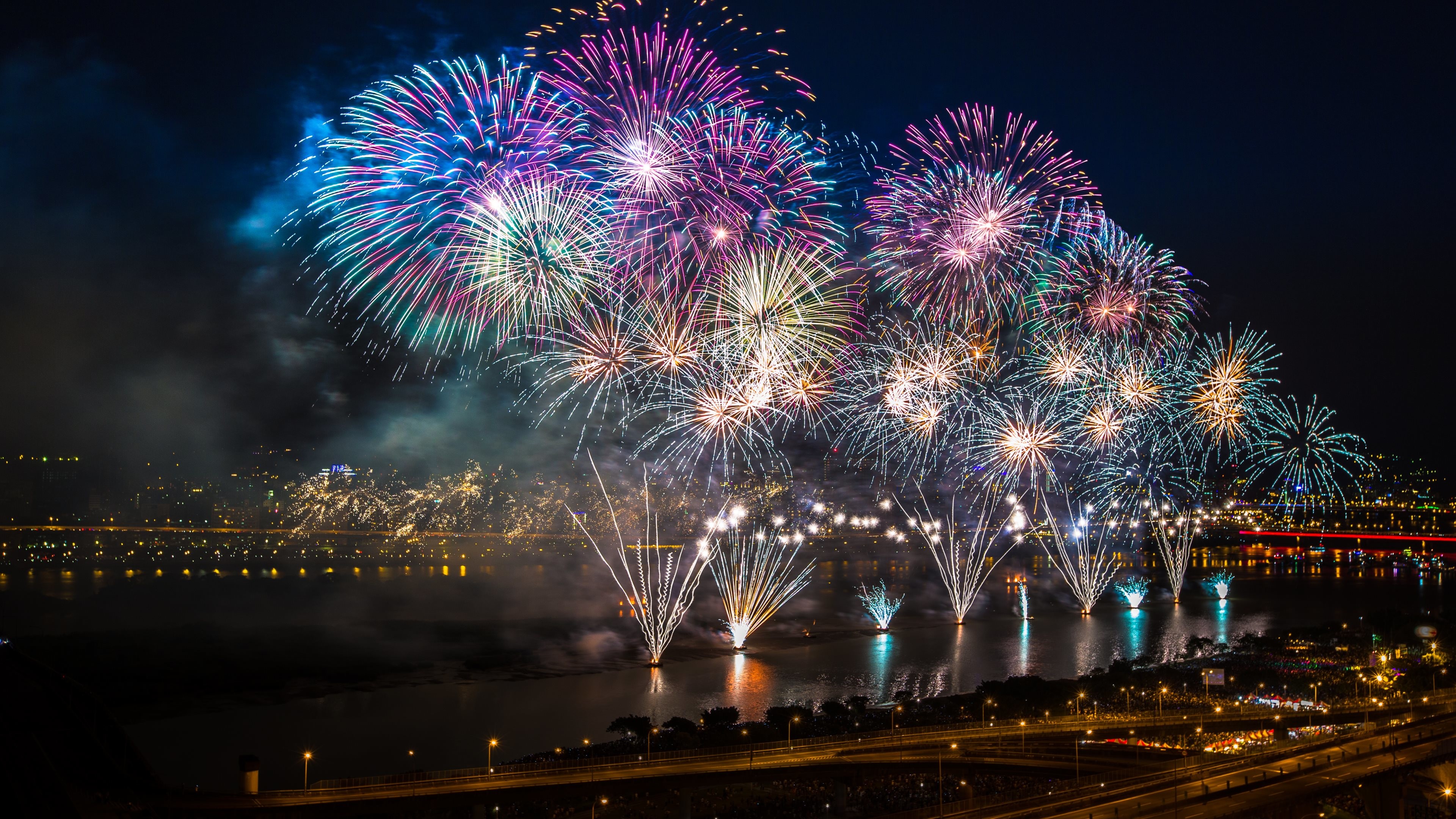 Fireworks light up the night sky over a city, reflecting in the water below, creating a spectacular New Year's Eve scene