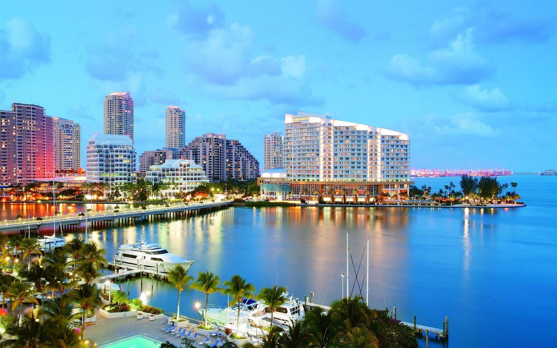 Scenic view of the Miami skyline at dusk, featuring illuminated buildings, calm waters, and docked boats