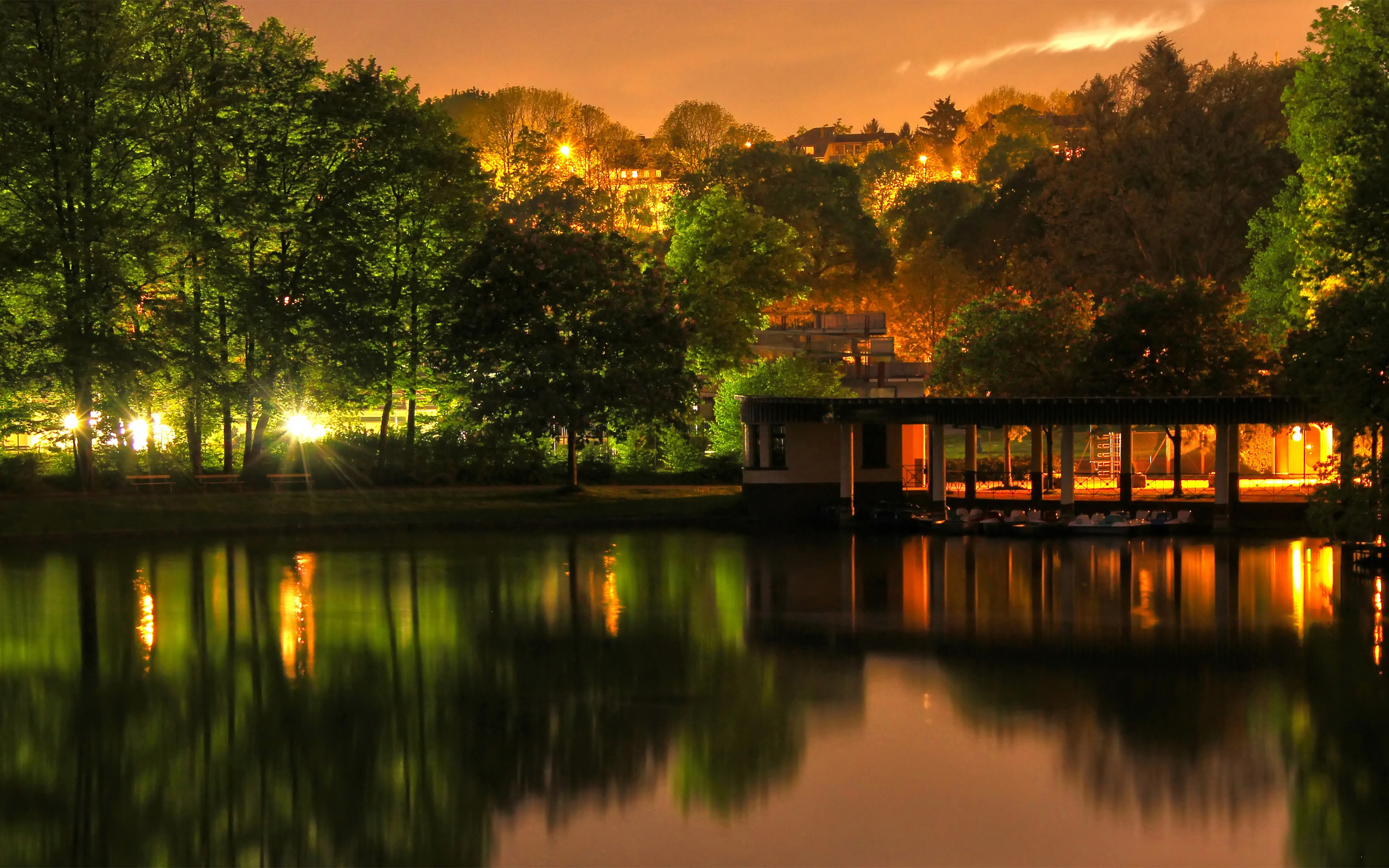 A tranquil lake reflecting the warm glow of lights at sunset, surrounded by lush trees and a charming boathouse