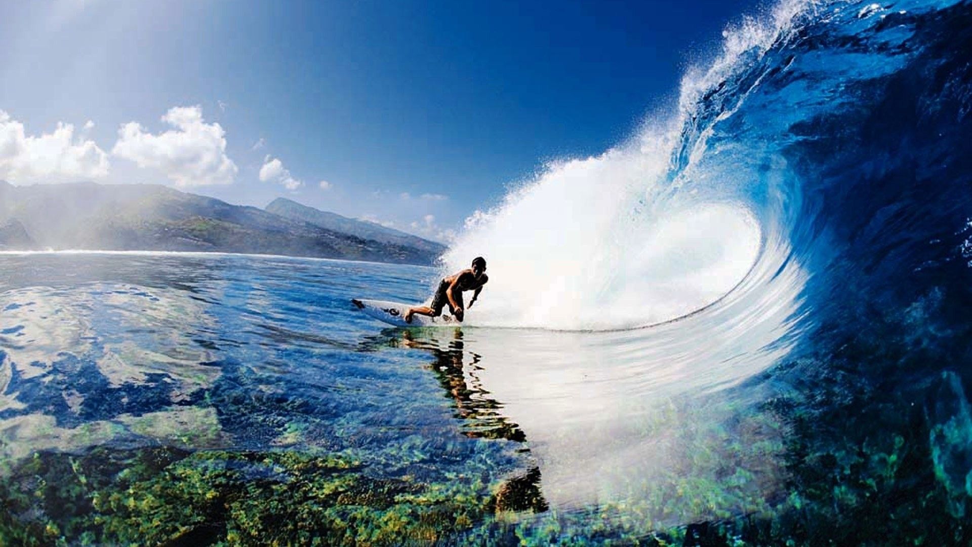 A surfer rides a crystal-clear wave with a backdrop of a tropical island and blue skies