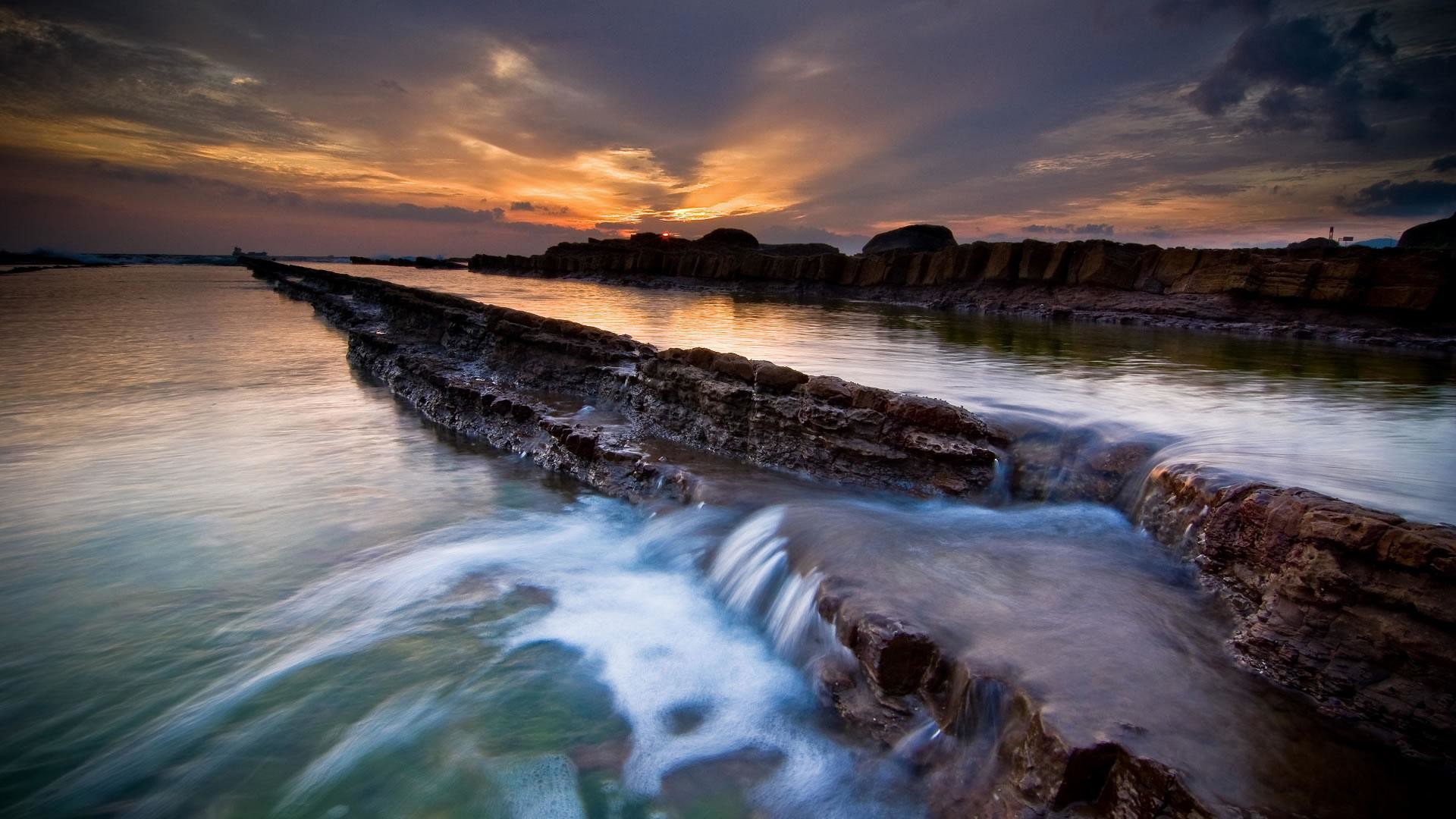 Stunning sunset over a rocky coastline with flowing water, perfect for a scenic computer wallpaper