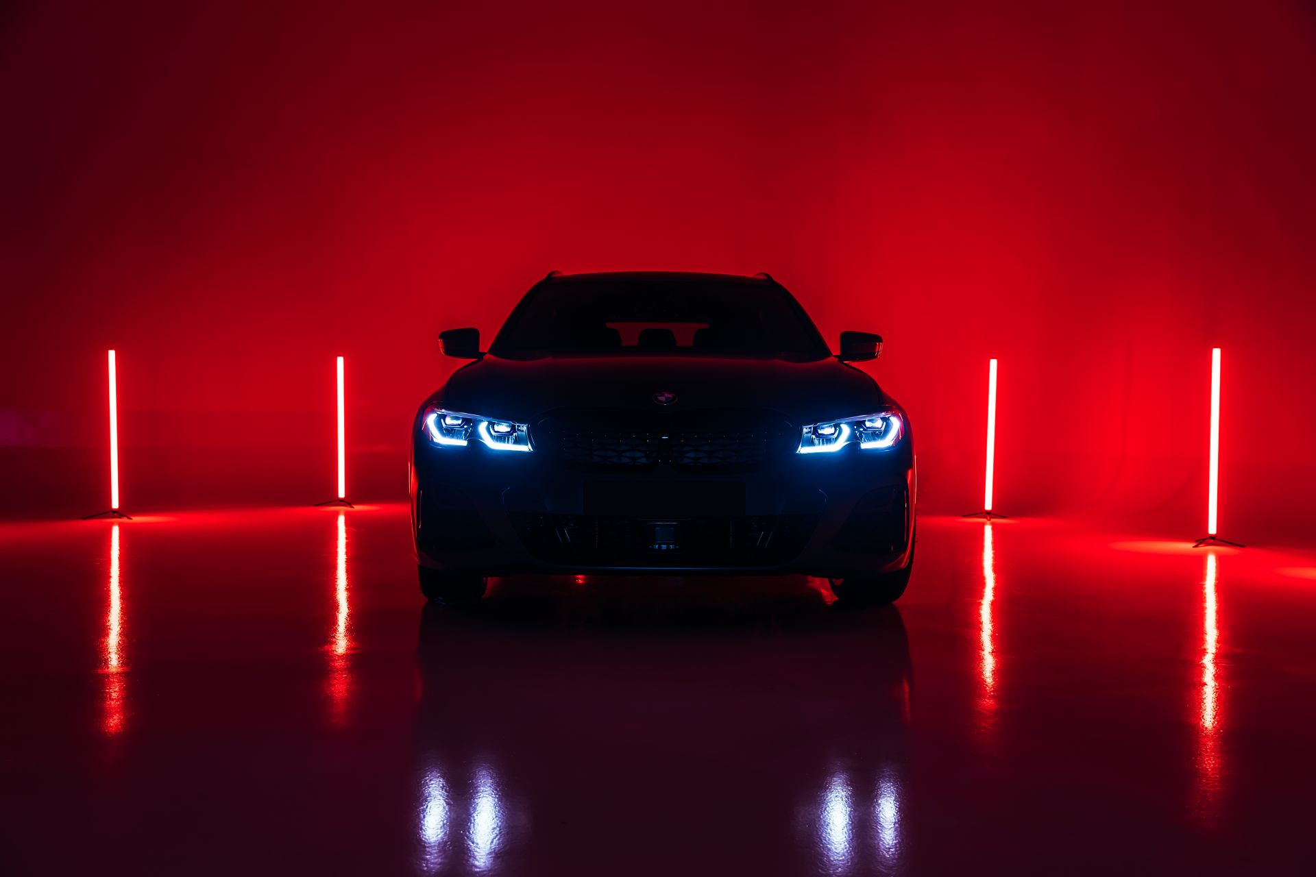 A dark BMW car with its headlights on, parked in a studio with a red background and neon lights
