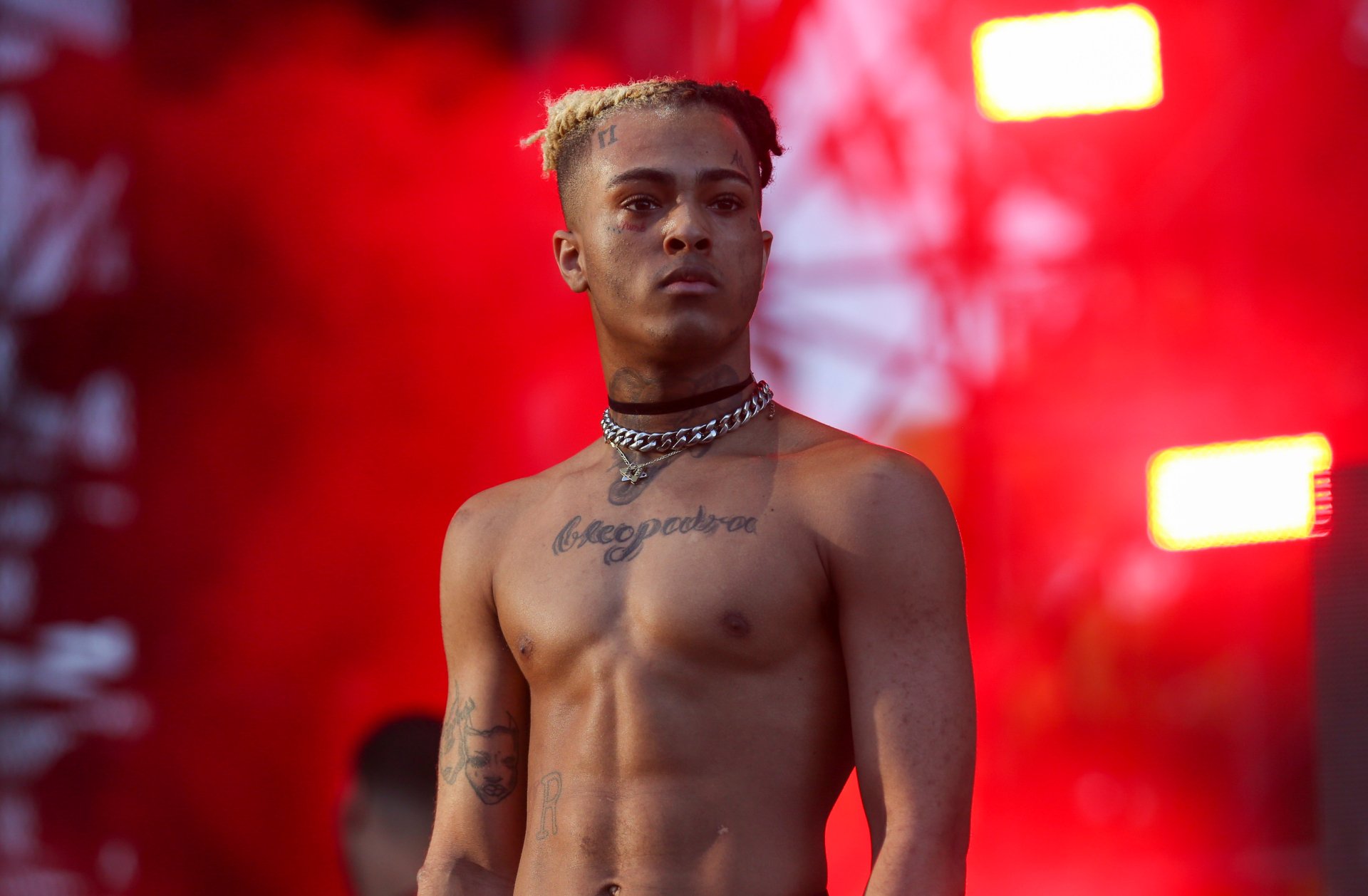 XXXTentacion performing live on stage, shirtless and displaying his tattoos. The background is a vibrant red, likely from stage lighting