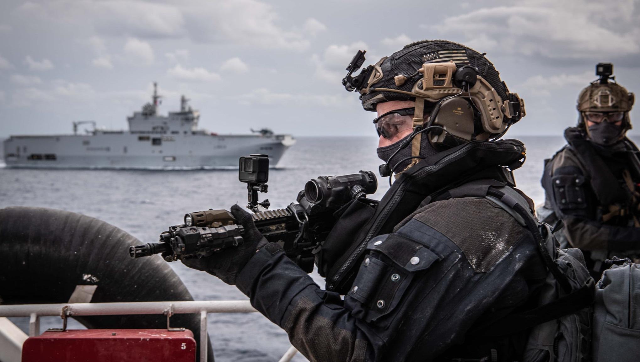 A French Commandos Marine soldier stands watch on a ship, assault rifle equipped with advanced optics, with a warship in the background