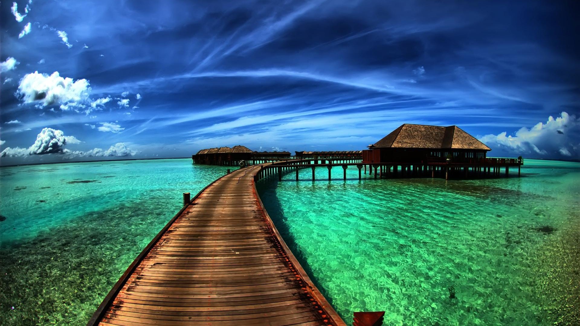 A wooden pier stretches towards overwater bungalows in crystal-clear turquoise water, under a vibrant blue sky dotted with fluffy white clouds