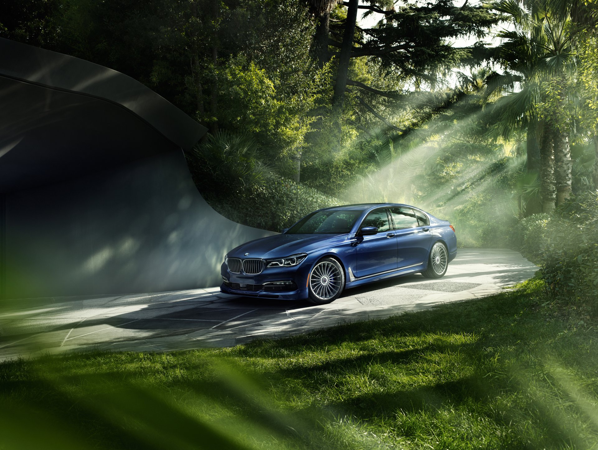 A blue BMW 7 Series sedan parked on a paved driveway surrounded by lush greenery