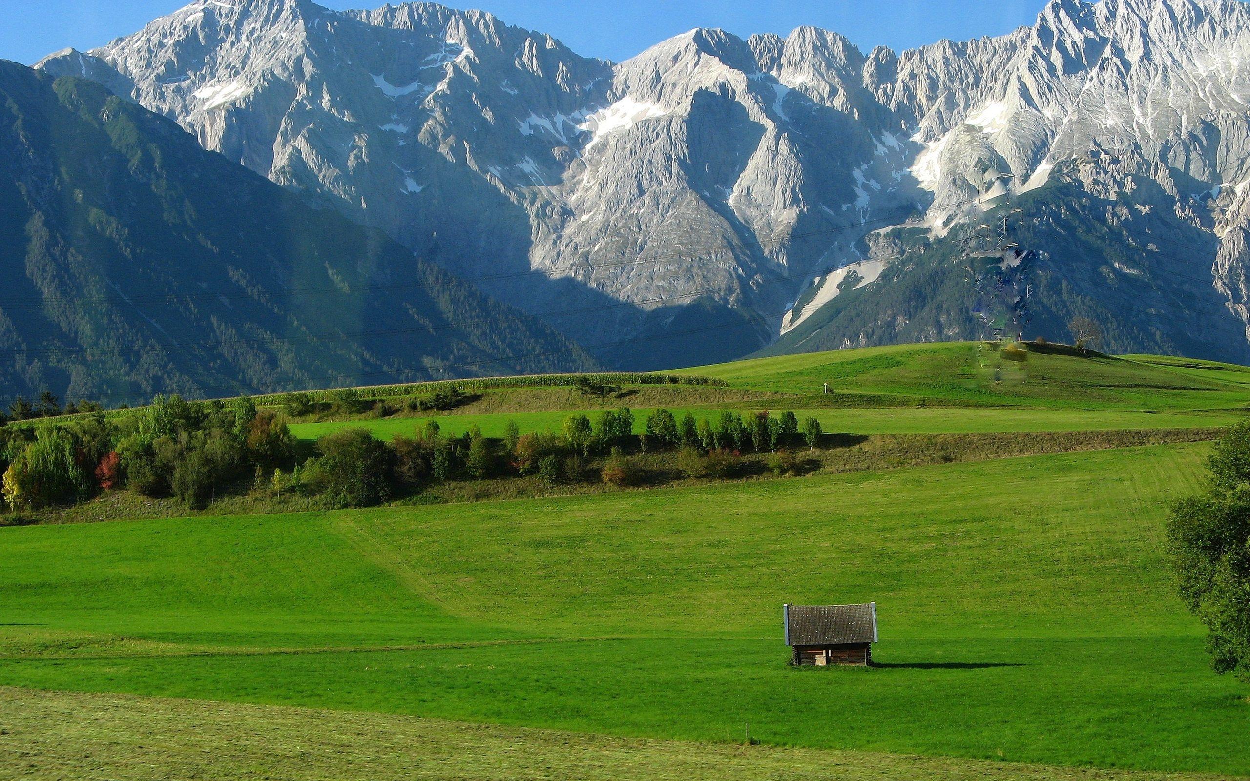 A breathtaking landscape of rolling green hills, dotted with trees and a small wooden hut, set against the majestic backdrop of snow-capped mountains