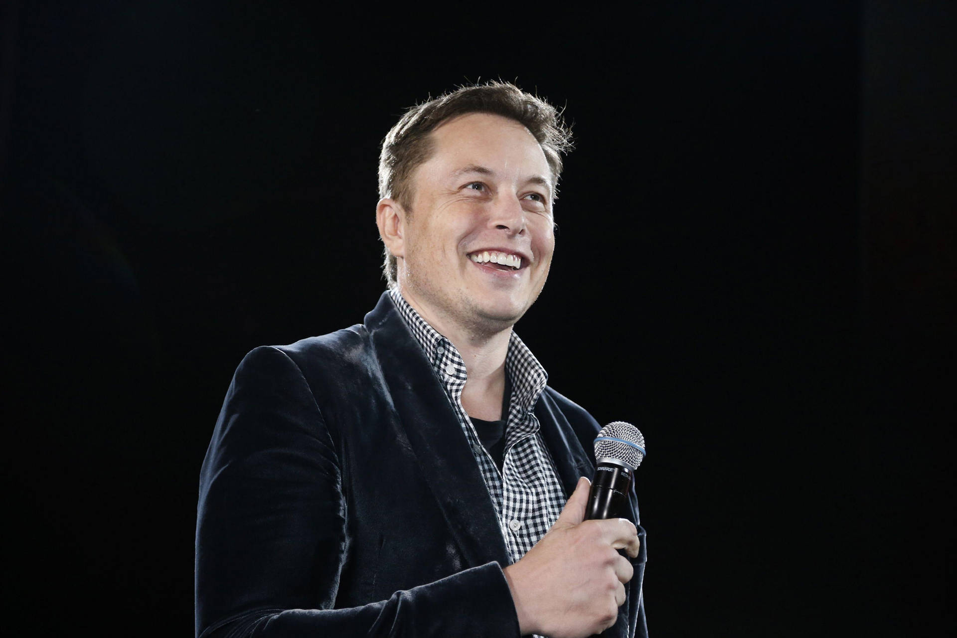 Elon Musk smiles at an event, holding a microphone
