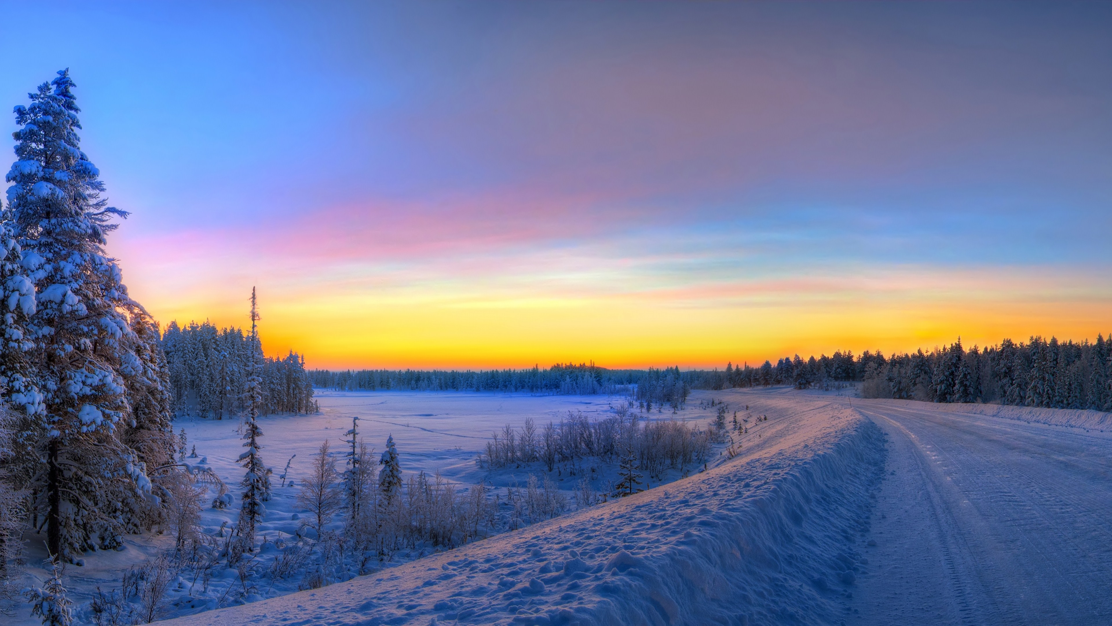 A breathtaking 4k winter wallpaper showcasing a snow-covered landscape at sunrise. The colorful sky blends from blue to pink and orange, illuminating the frosty trees and the pristine snow. A road winds through the scene, inviting viewers to experience the tranquil beauty of winter