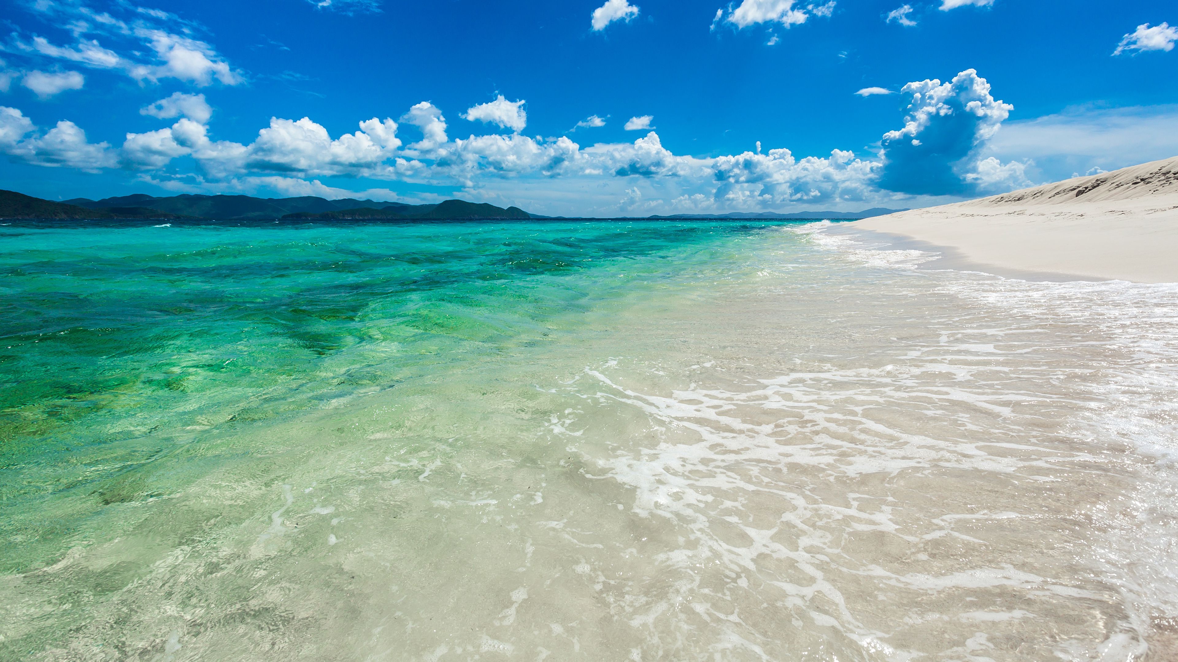 Crystal-clear turquoise water gently laps against a pristine white sand beach, with lush green hills in the distance under a vibrant blue sky dotted with fluffy white clouds