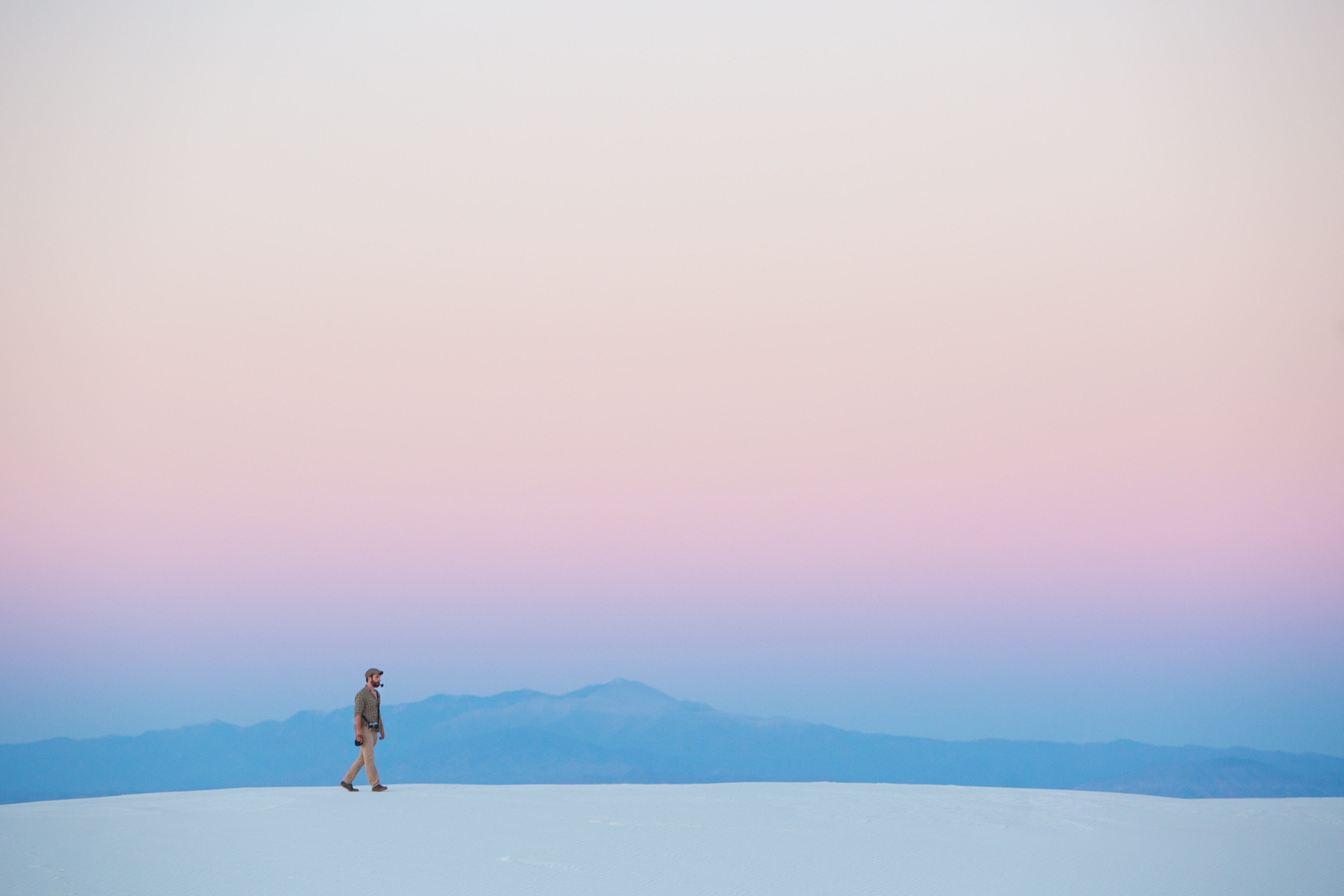A minimalist landscape photograph of a lone figure walking on a white sand dune against a pastel sunset sky with distant blue mountains