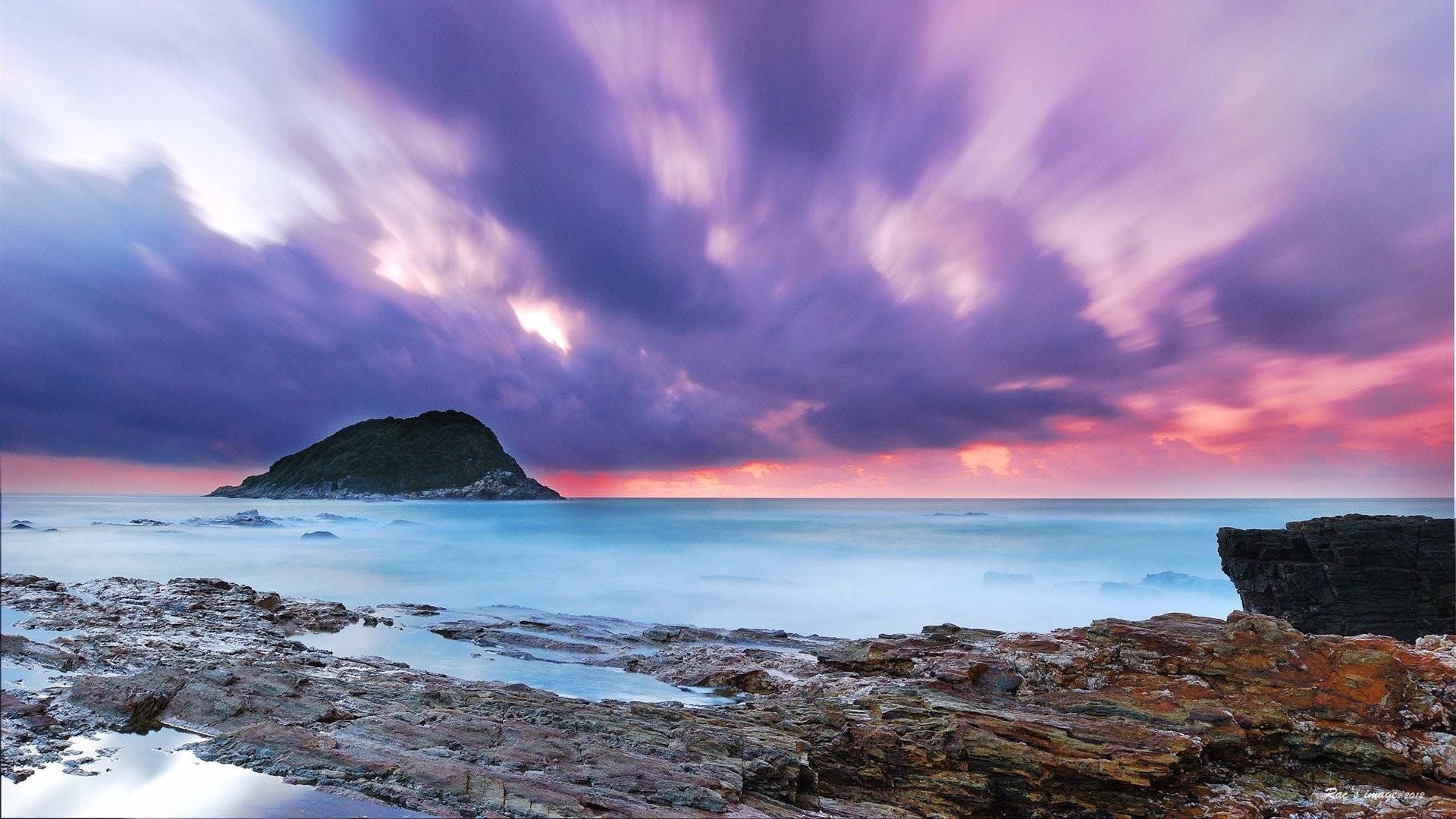 A breathtaking sunrise paints the sky in vibrant hues of purple and pink over a tranquil ocean, with a rocky coastline in the foreground and a small island silhouetted against the colorful horizon
