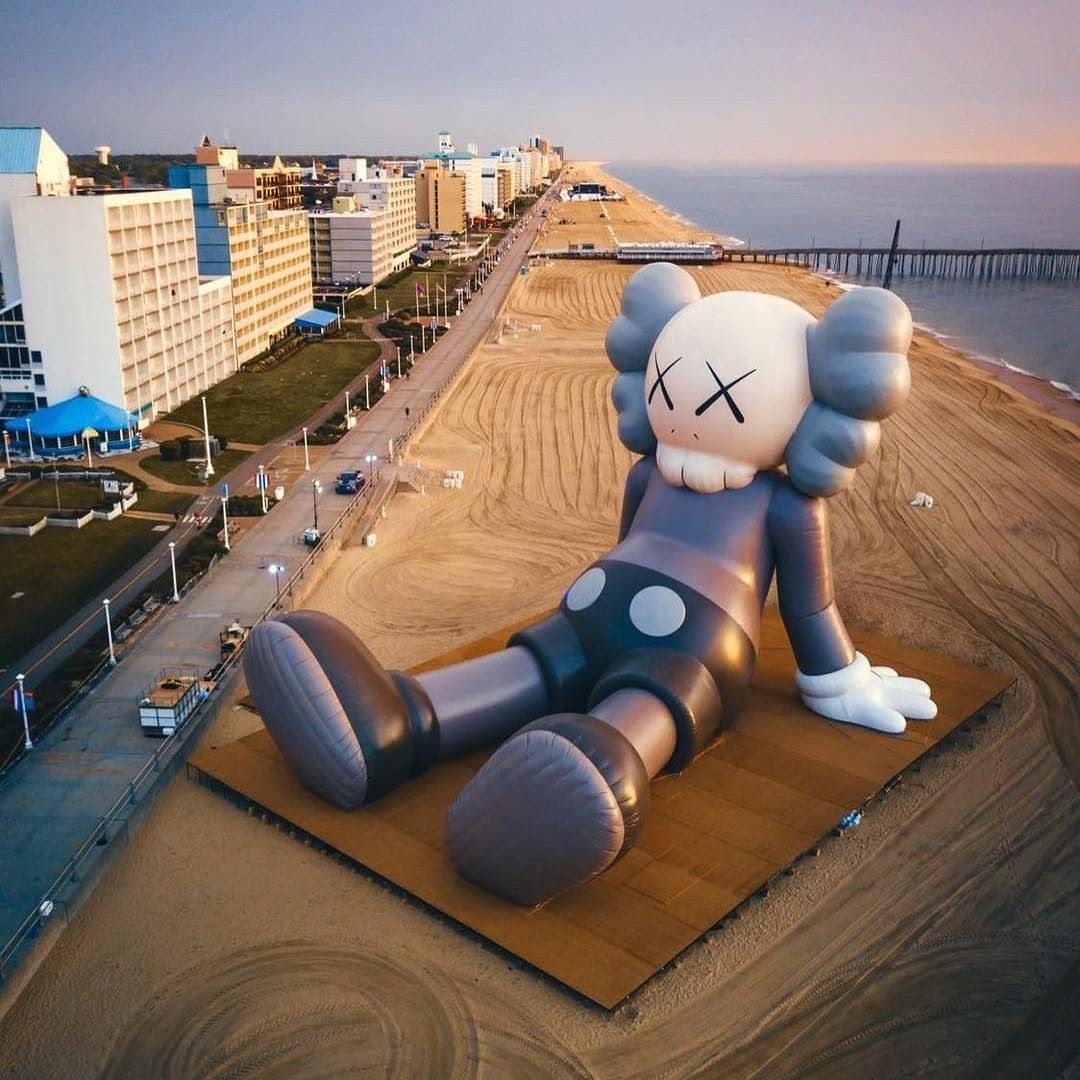 A giant inflatable KAWS Companion figure rests on a wooden platform on a sandy beach, with the ocean and a coastal city in the background