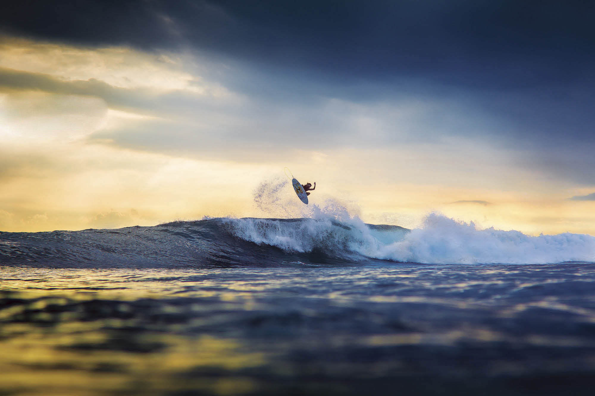 A surfer performs an aerial maneuver on a wave at sunset, silhouetted against the golden sky
