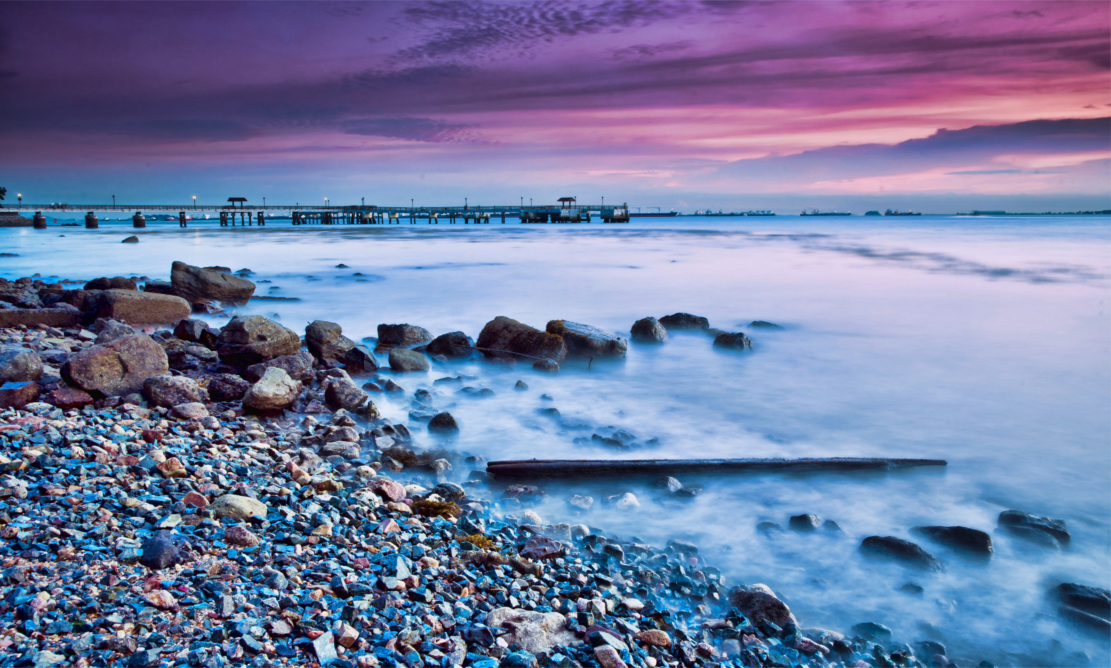 A serene long exposure photo of a rocky beach at sunset, with a pier in the background and a purple and pink sky