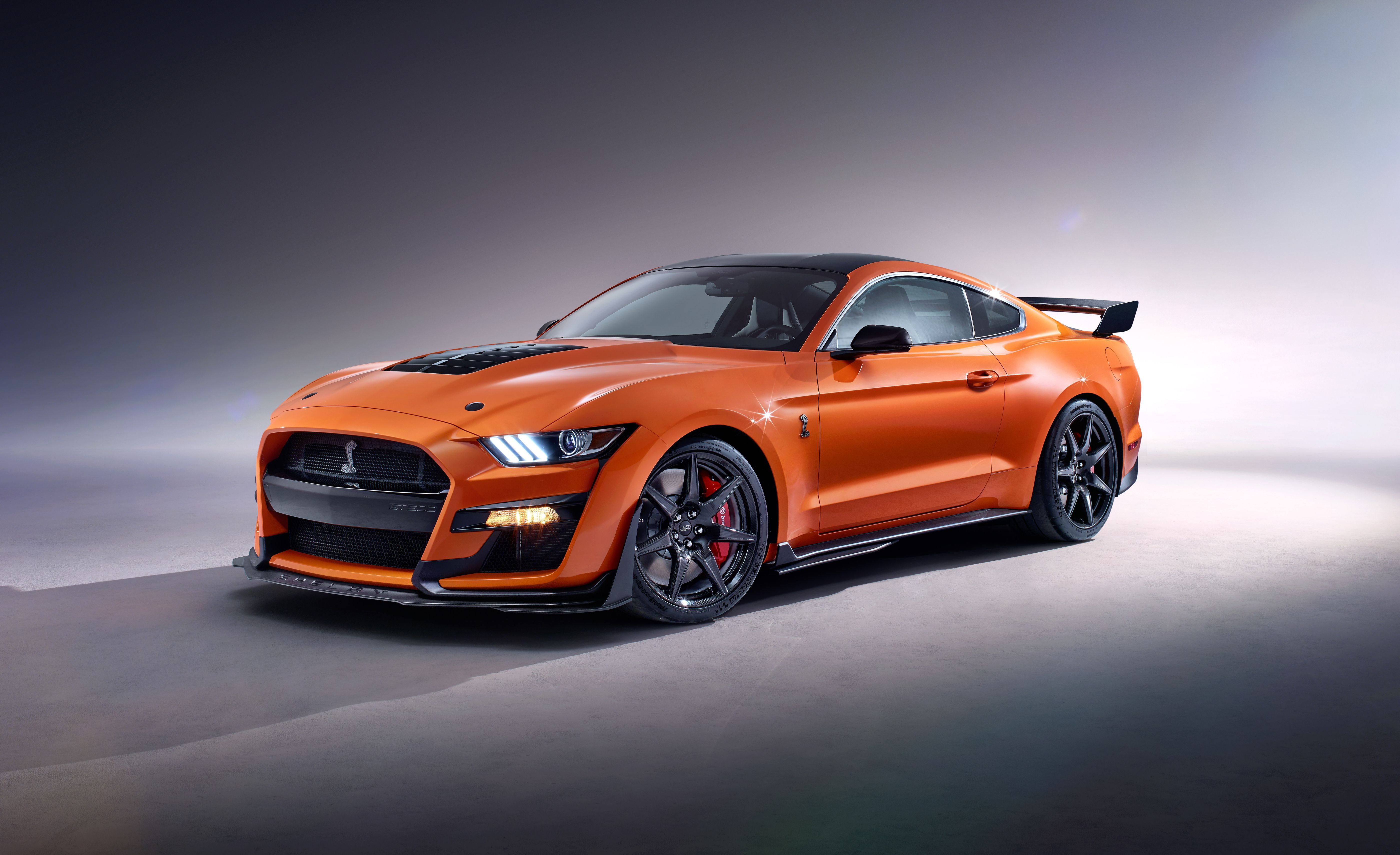 A vibrant orange Ford Mustang Shelby GT500, parked and gleaming under studio lighting. This iconic muscle car showcases its aggressive lines, hood vents, and rear spoiler, emphasizing its powerful presence