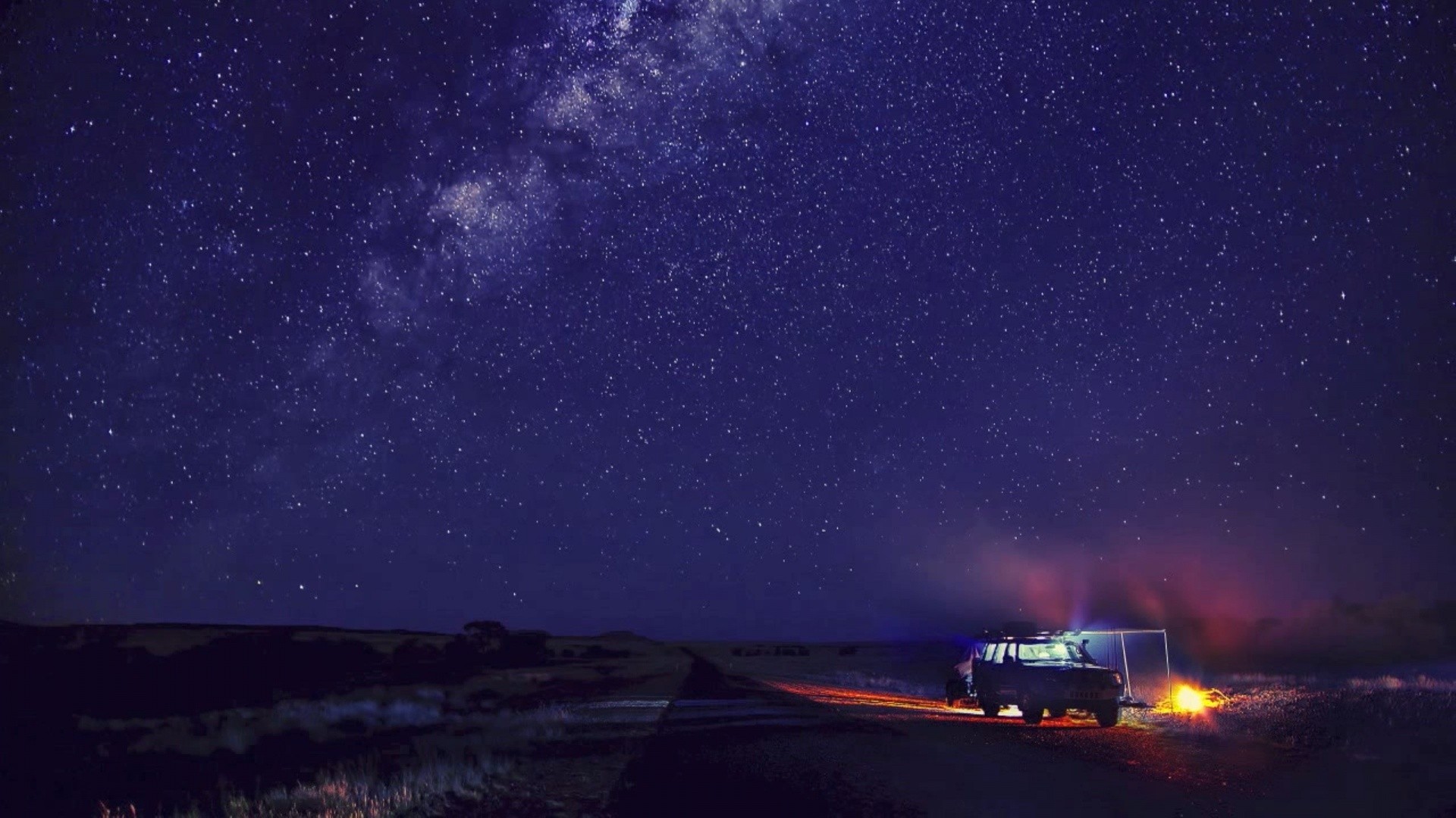 A breathtaking 4K wallpaper of the night sky, showcasing the Milky Way over a serene landscape. A vehicle is parked near a campfire, illuminated by the starry expanse