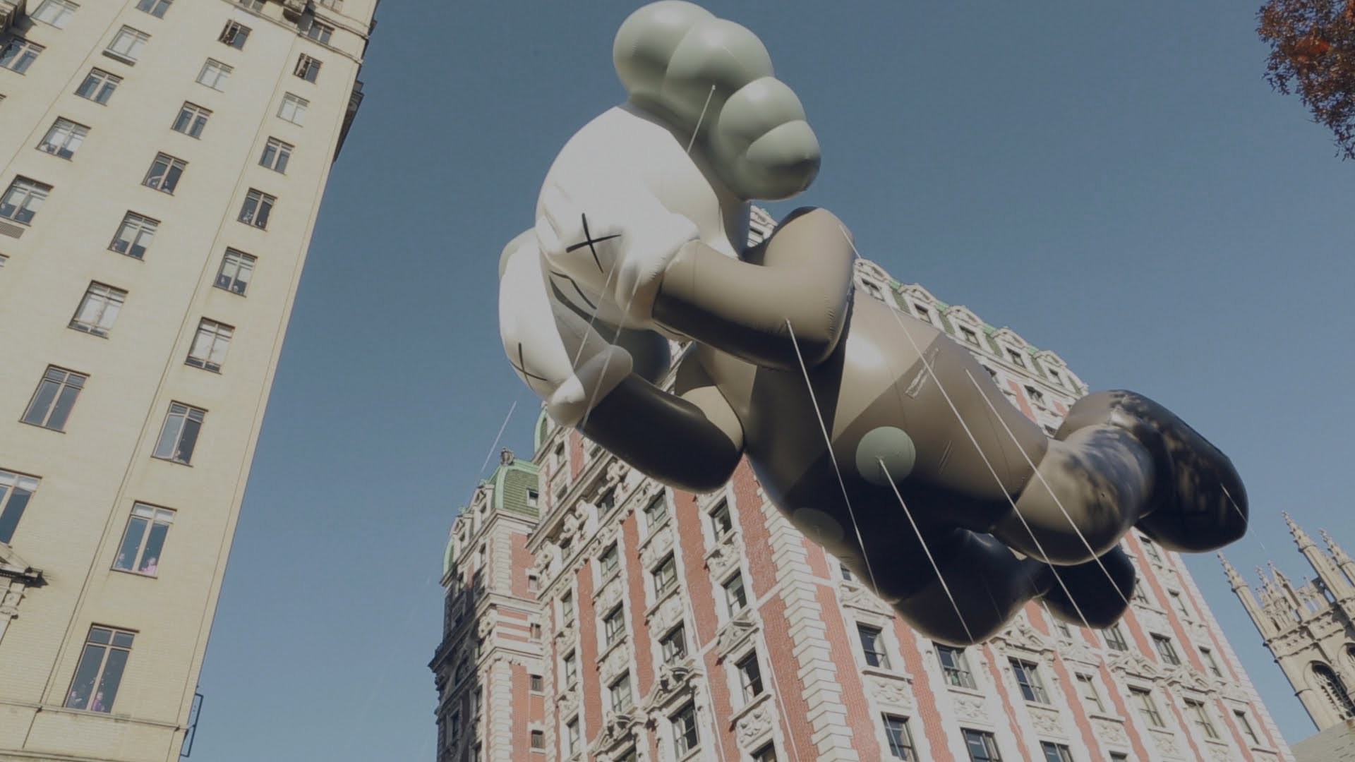 A low-angle view of a KAWS Companion inflatable balloon floating between buildings, likely during a parade