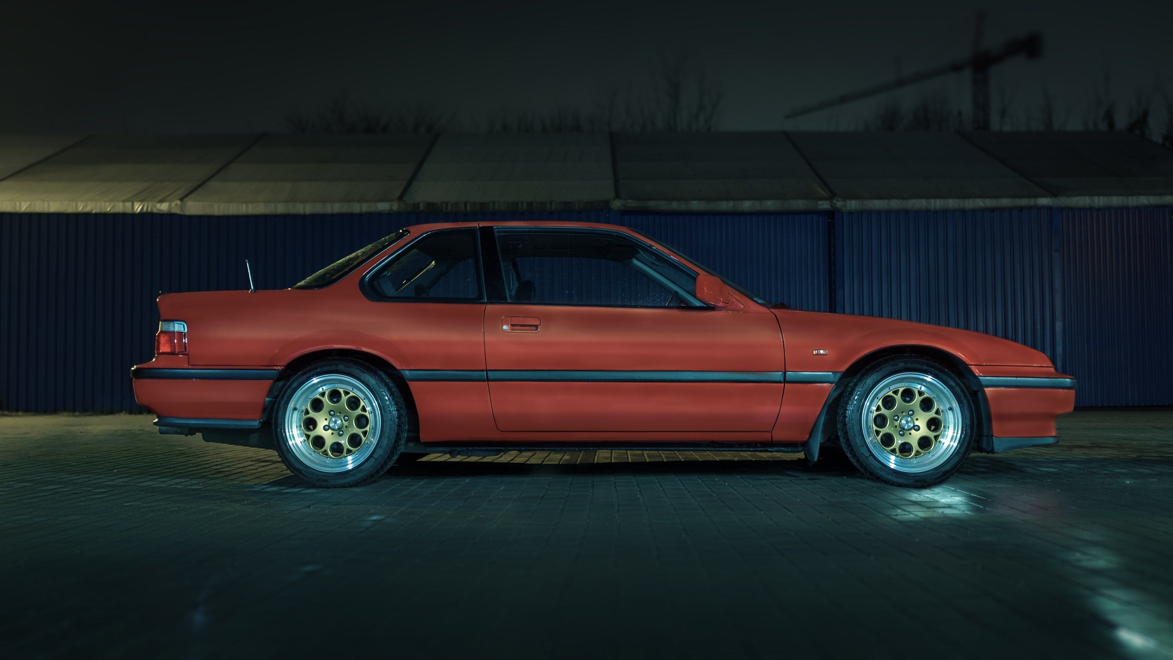 A red Honda Prelude parked in front of a blue fence at night, showcasing its sleek profile and gold rims