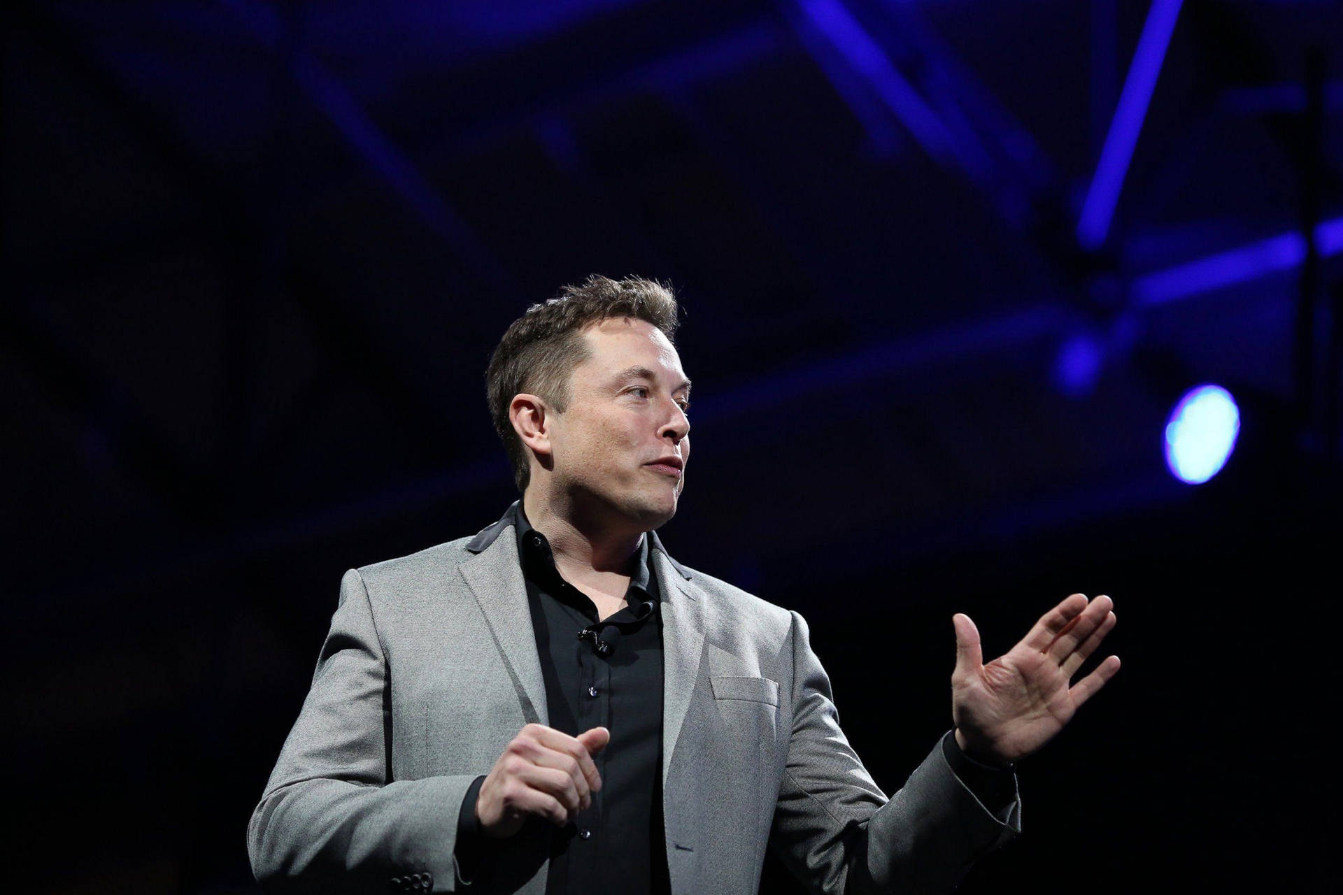 Elon Musk speaking at a conference, wearing a gray suit and black shirt