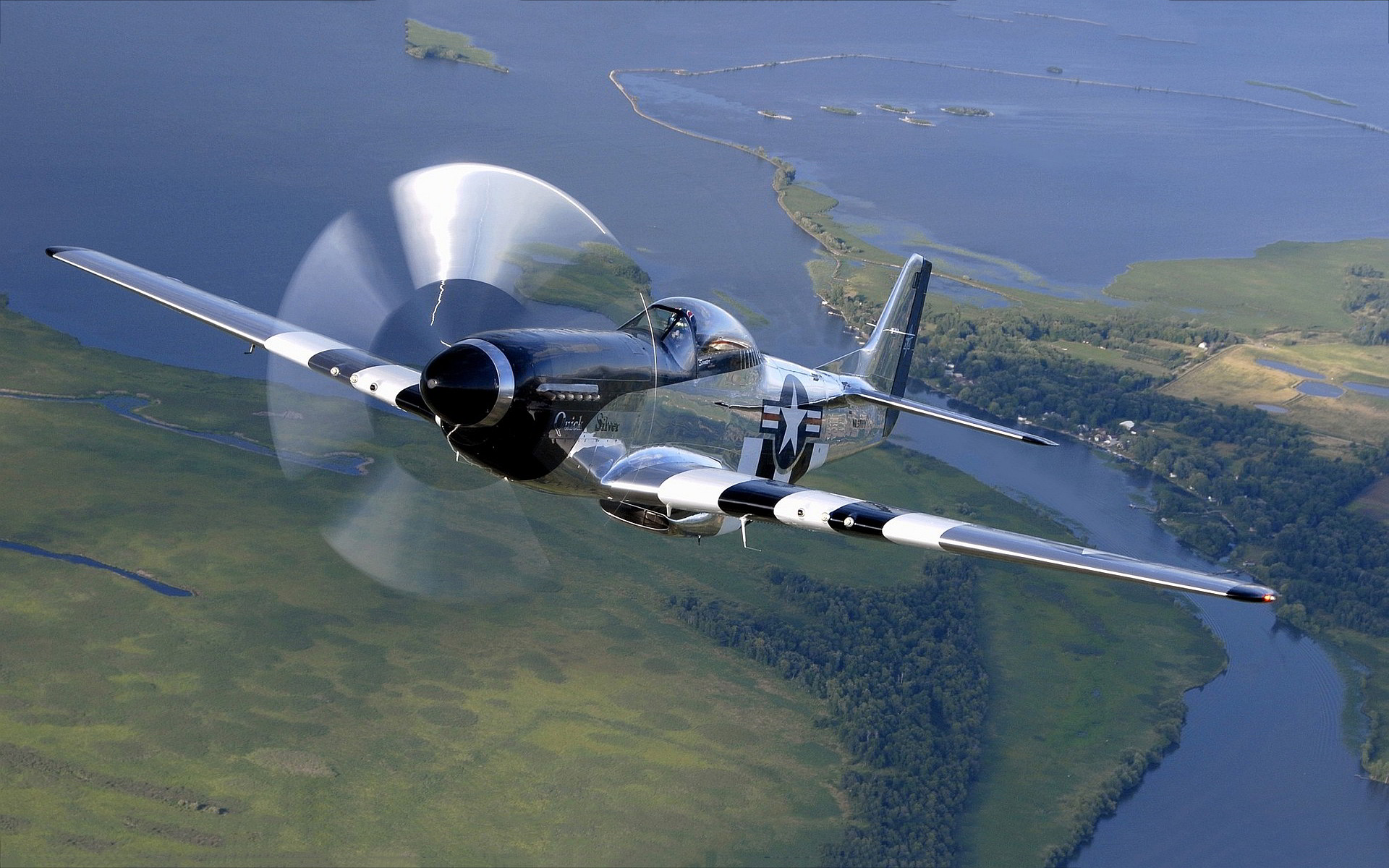 A silver and black P-51 Mustang fighter plane in flight over a landscape of green and blue